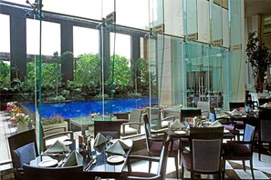 An indoor dining area with tables and chairs, a glass wall overlooking a garden and a pool.