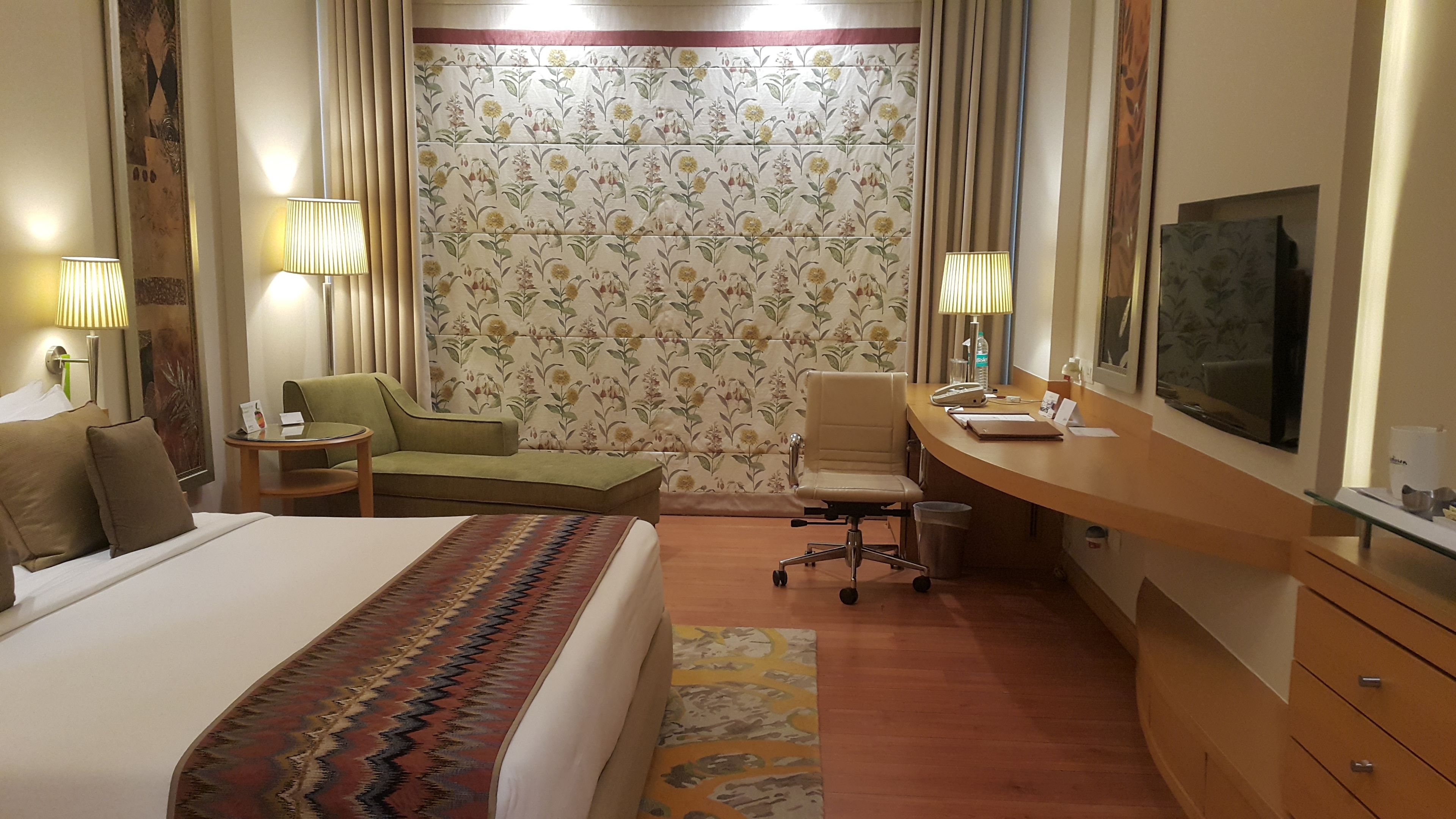 A hotel room with a bed, a desk, a chair, a small table, a trash bin, and a large mirror.