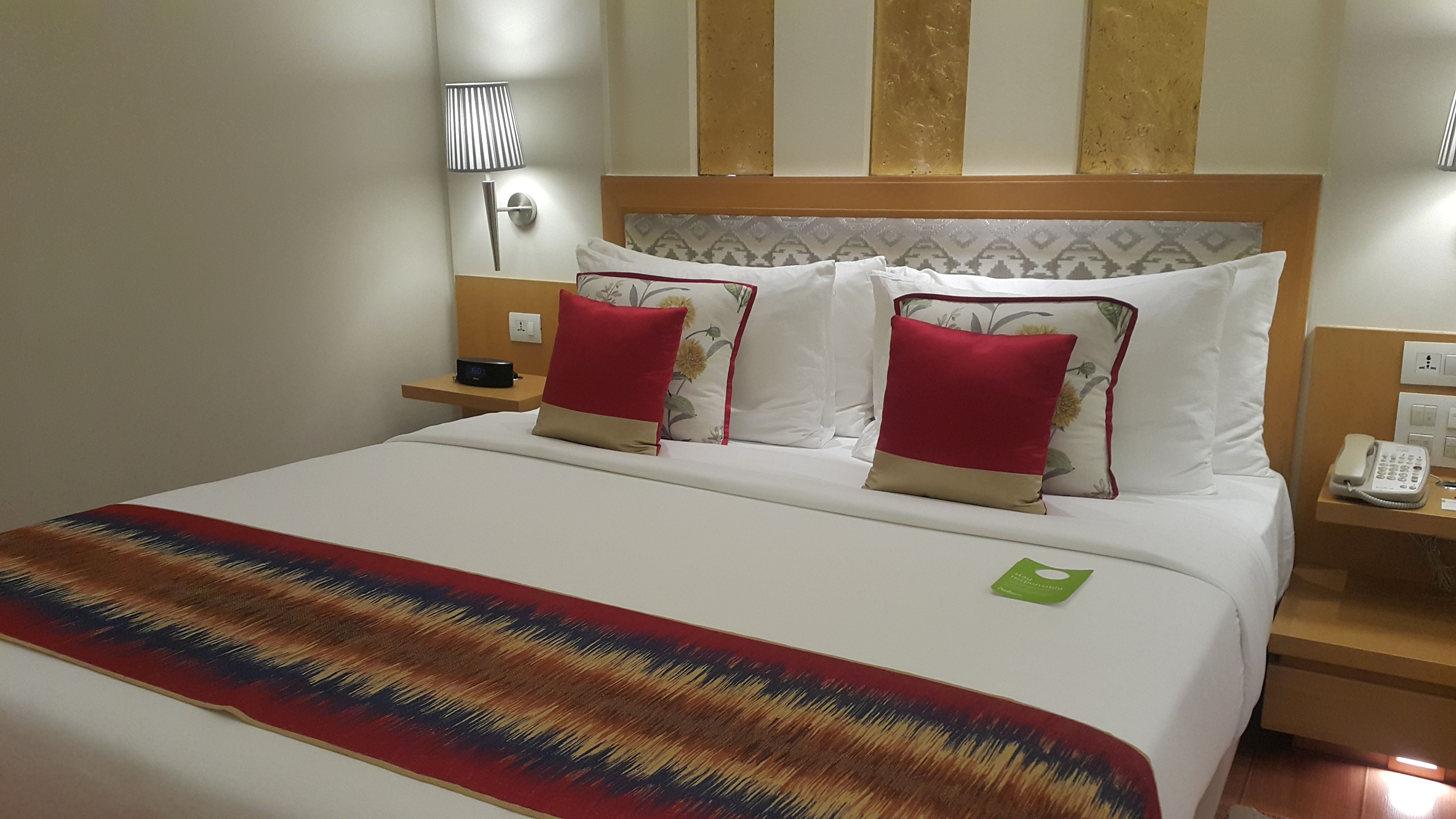 A hotel room with a large bed, two red and beige pillows, a bedside table with a phone, and a wall-mounted lamp.