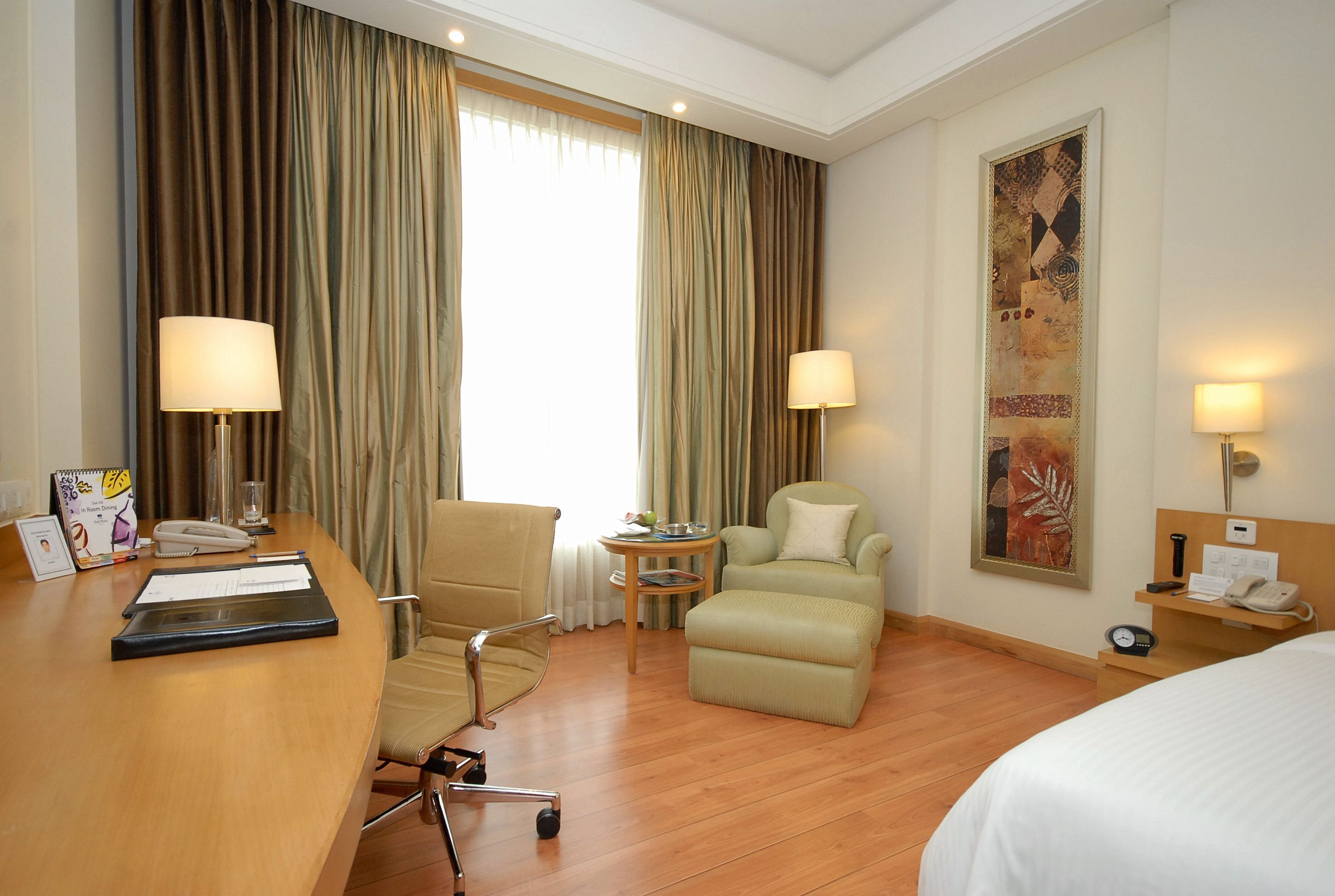 A hotel room with a wooden floor, a desk with a lamp, a chair, a sofa, a bedside table with a lamp, and a large painting on the wall.