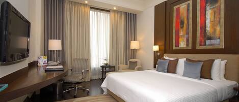 A hotel room with a large bed, a desk with a flat-screen TV, a chair, a small table with a lamp, and two paintings on the wall.
