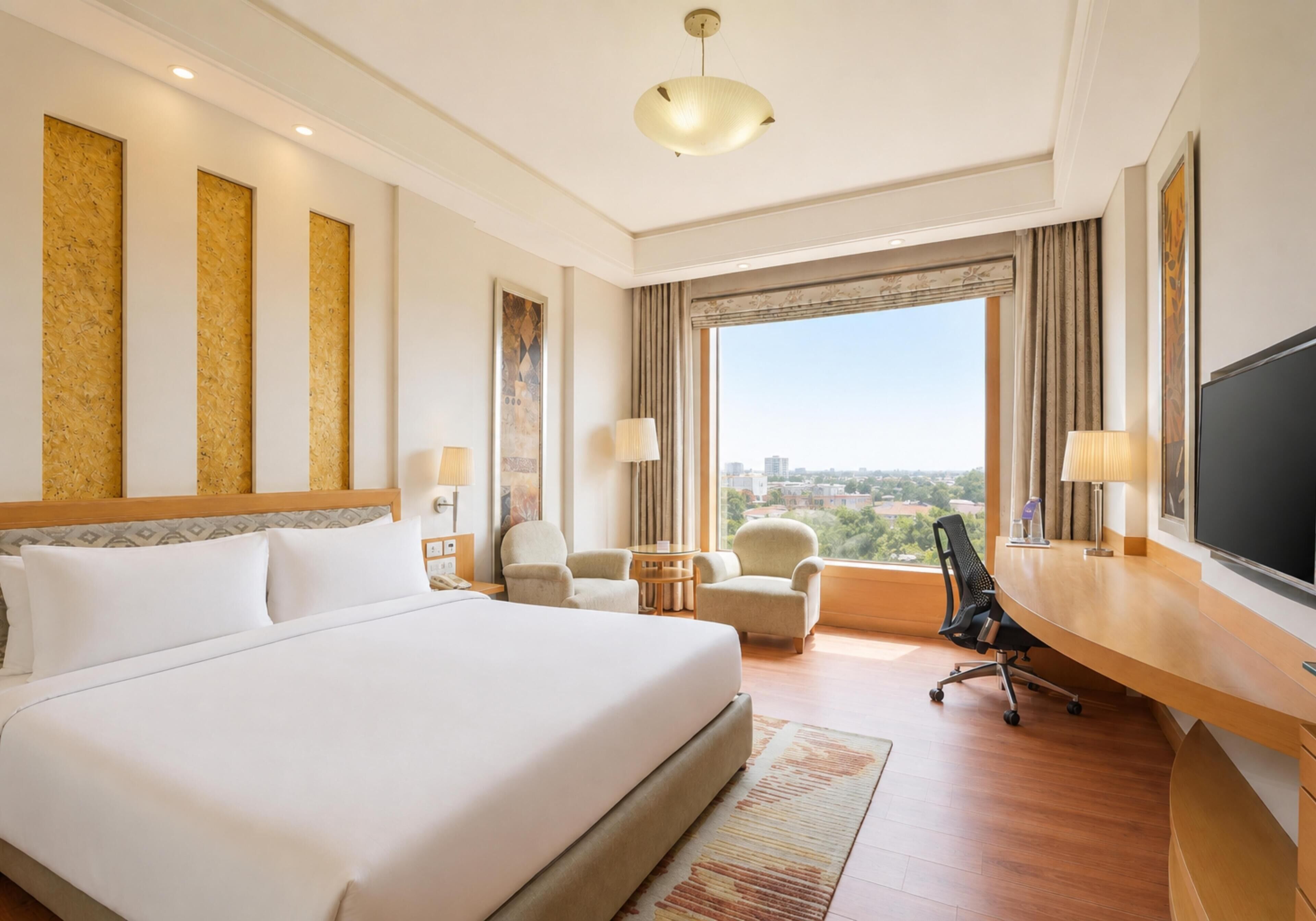 A hotel room with a large bed, two chairs, a desk, a television, and a window with a city view.