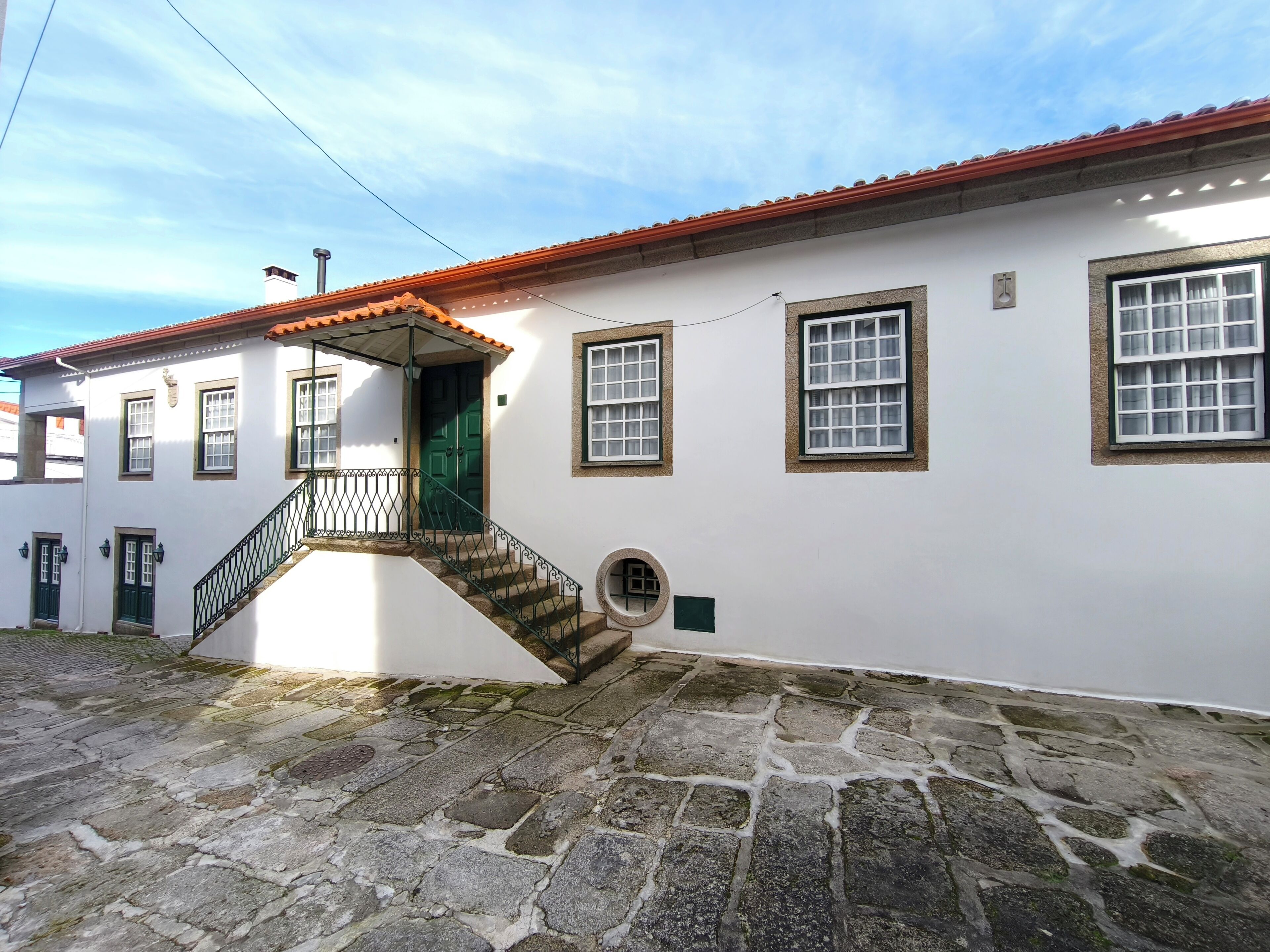 A white building with a red-tiled roof, a stone staircase, and multiple windows with green shutters.