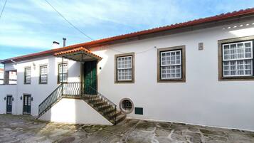 A white building with a red-tiled roof, a stone staircase, and multiple windows with green shutters.