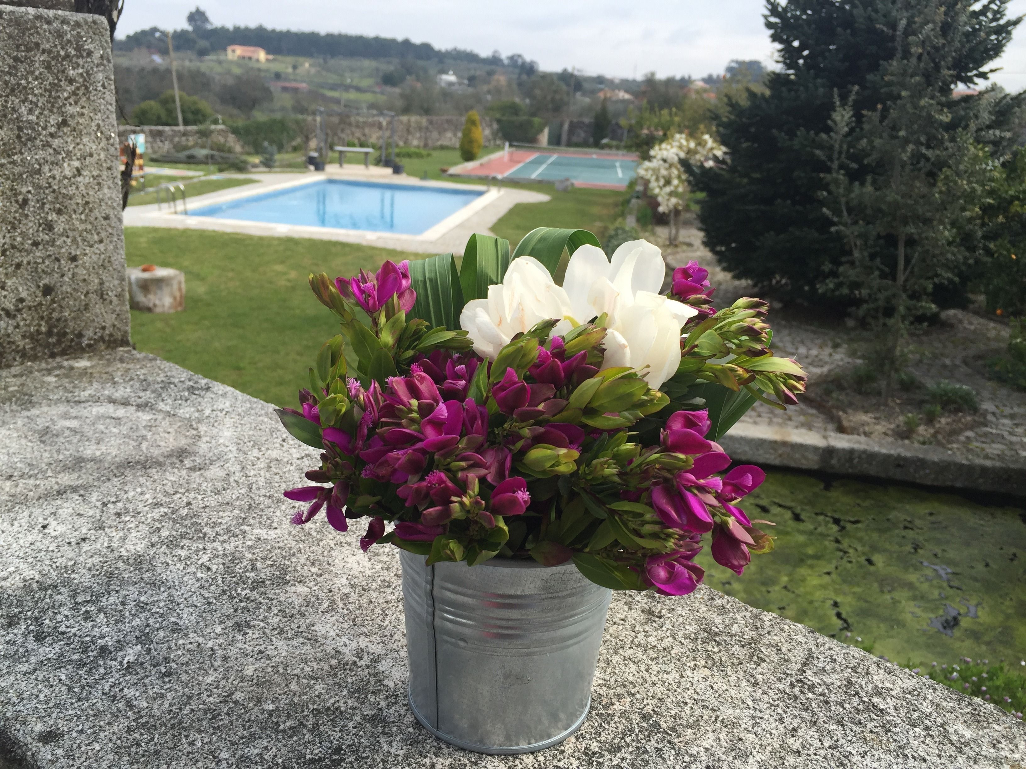 A flower arrangement in a metal pot on a stone ledge with a view of a swimming pool and tennis courts in the background.