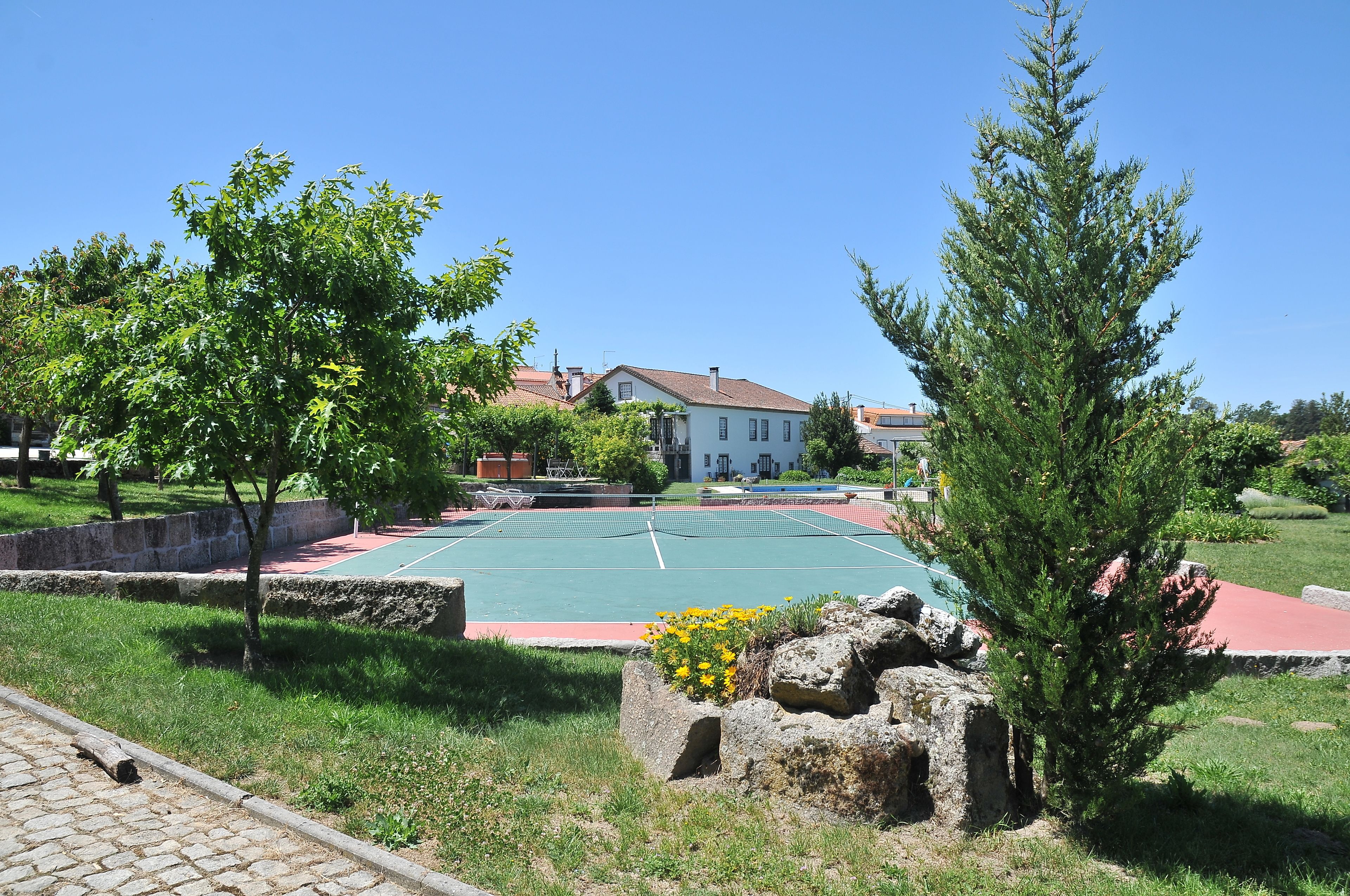 A tennis court surrounded by greenery and a stone wall, with a building in the background.