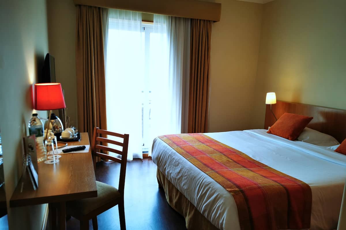 A hotel room with a bed, a desk with a chair, a TV, and a window with curtains.