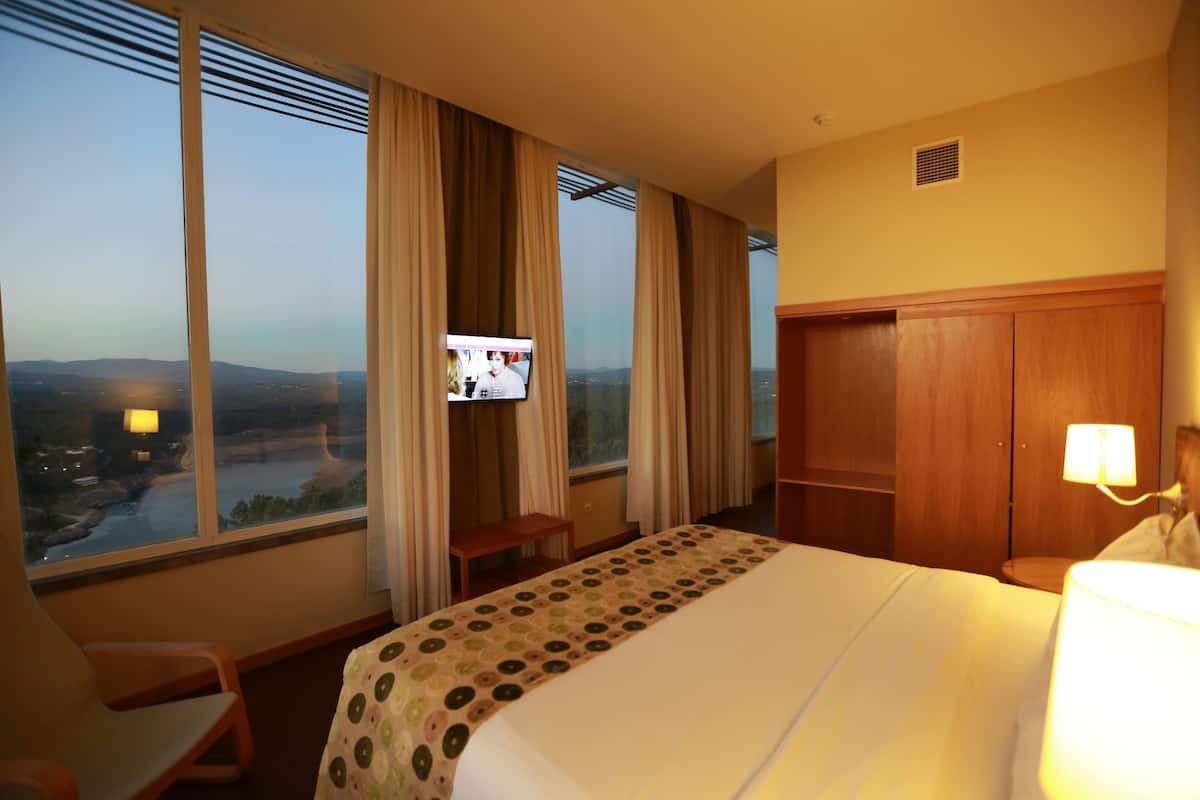 A hotel room with a bed, a desk, a chair, a TV, and a view of a cityscape.