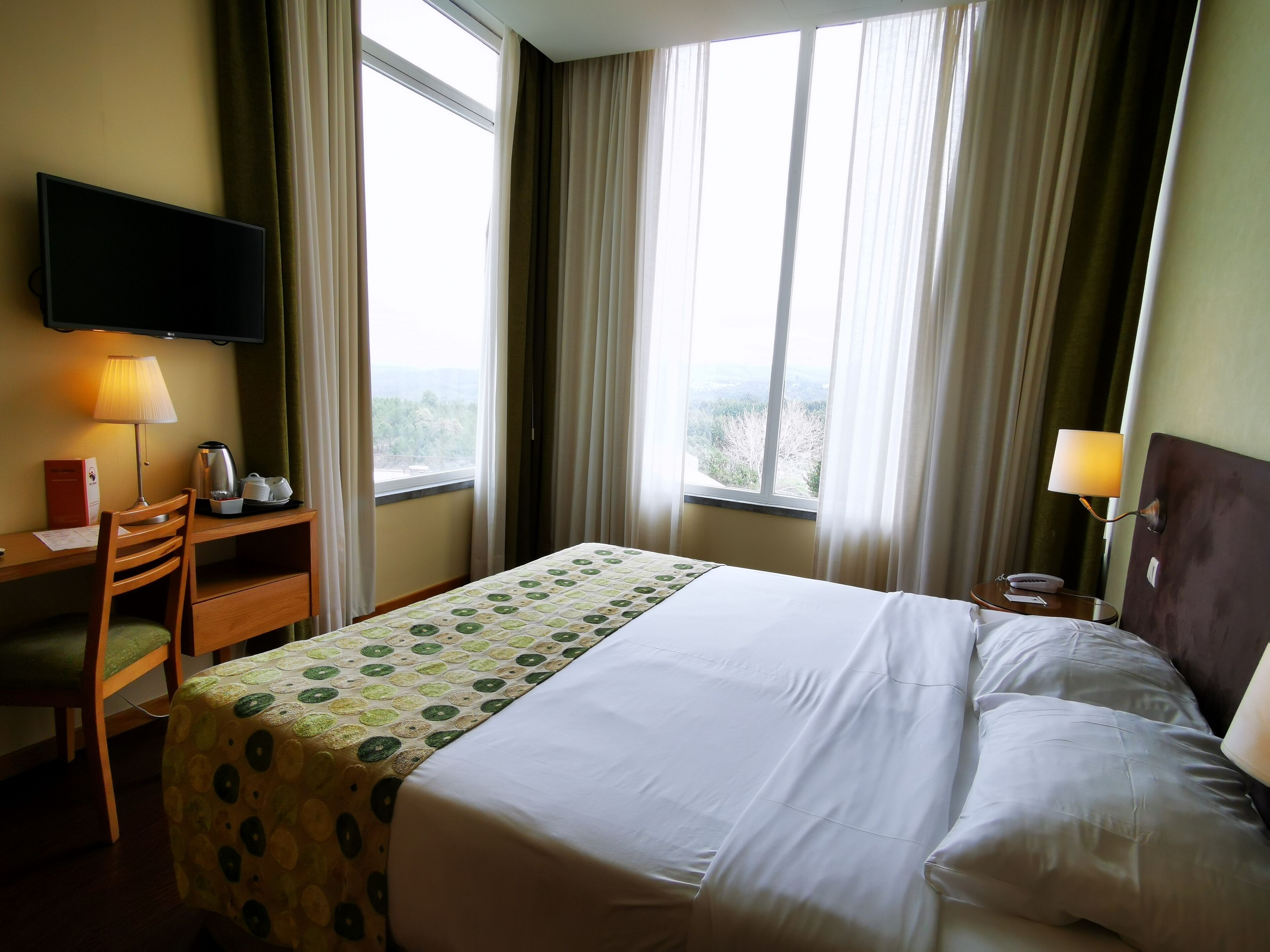 A hotel room with a bed, a desk with a lamp, a chair, a TV mounted on the wall, and a window with curtains.