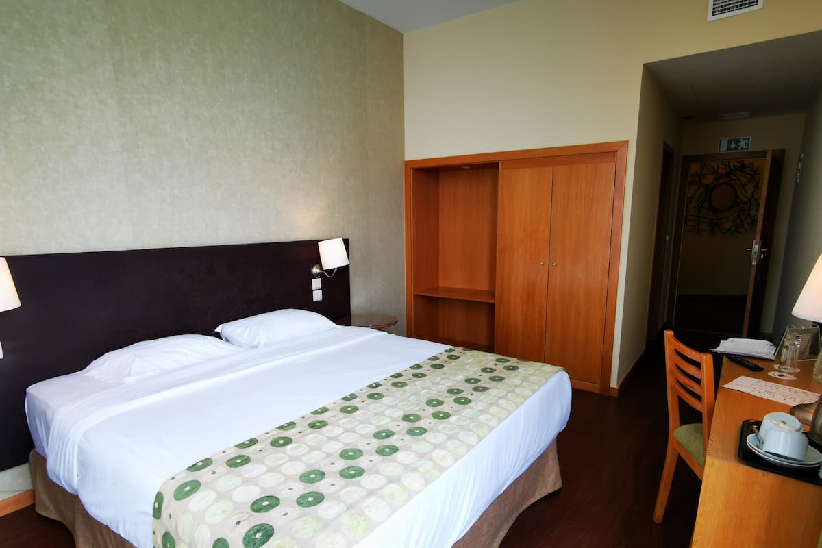 A hotel room with a bed, a desk with a chair, a wardrobe, and a lamp.