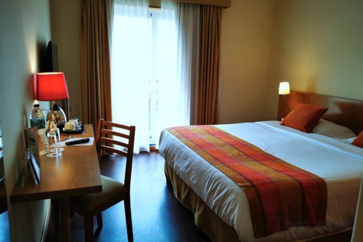 Habitación de hotel con cama, escritorio, silla, lámpara y vistas al exterior.