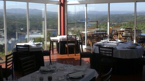 Un restaurante con amplios ventanales que ofrece una vista panorámica de montañas y un lago. Las mesas están dispuestas con copas de vino y platos.