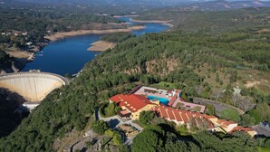 Vista aérea de un hotel con un edificio de techo rojo rodeado de un bosque denso, un gran embalse y montañas distantes.
