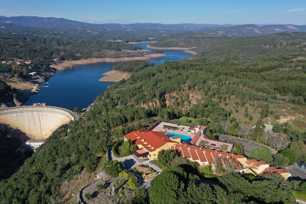Vista aérea de un hotel con un edificio de techo rojo rodeado de un bosque denso, un gran embalse y montañas distantes.