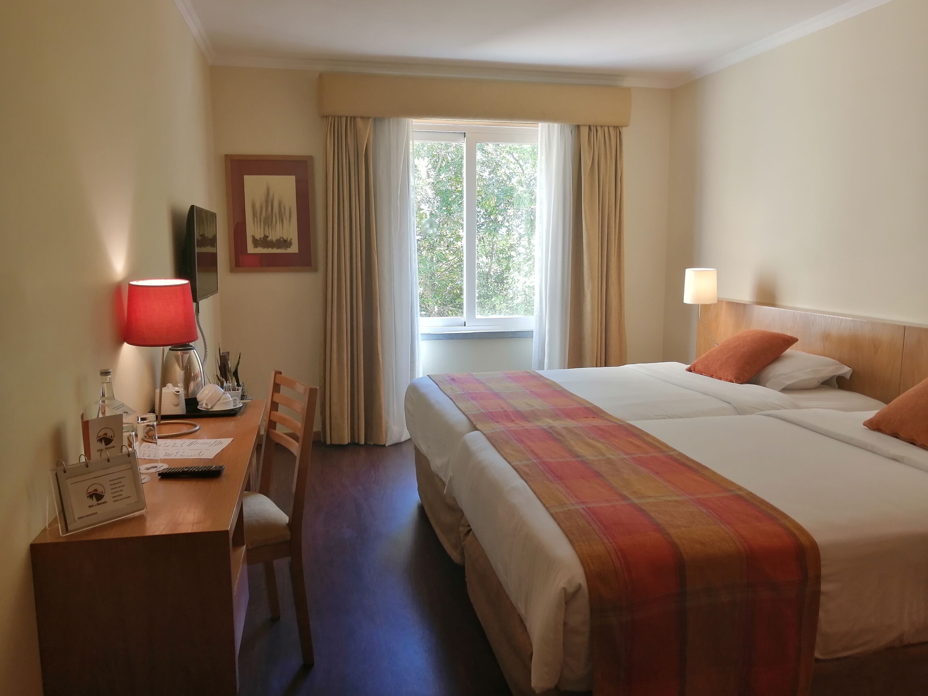 Habitación de hotel con cama, escritorio, silla, lámpara, ventana con cortinas e imagen enmarcada en la pared.