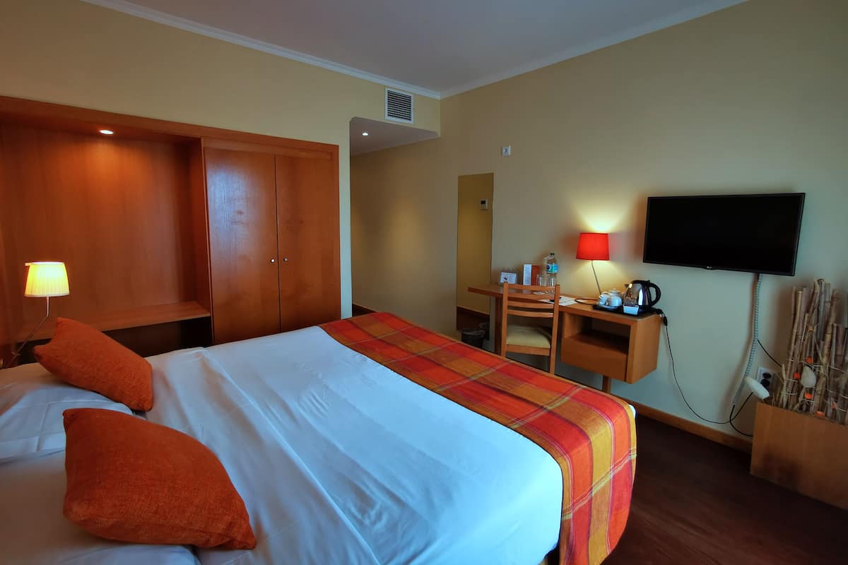 A hotel room with a bed, a desk, a chair, a television, and a small table with a kettle and cups.