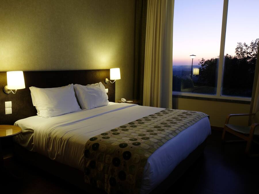 A hotel room with a large bed, two bedside lamps, a round table, and a view of the outdoors through a window.