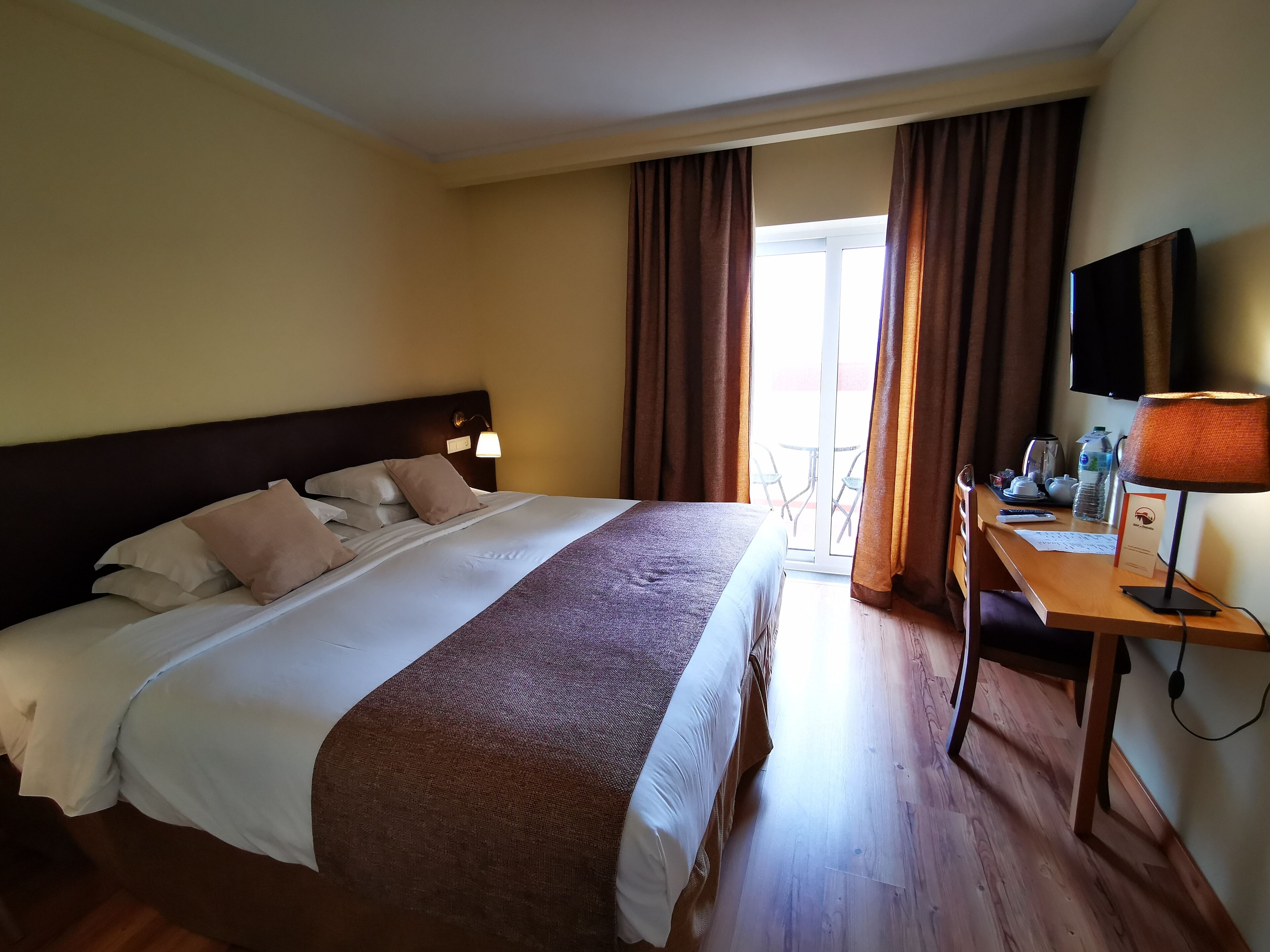 A hotel room with a large bed, a desk with a lamp, a television, and a view of the outdoors.