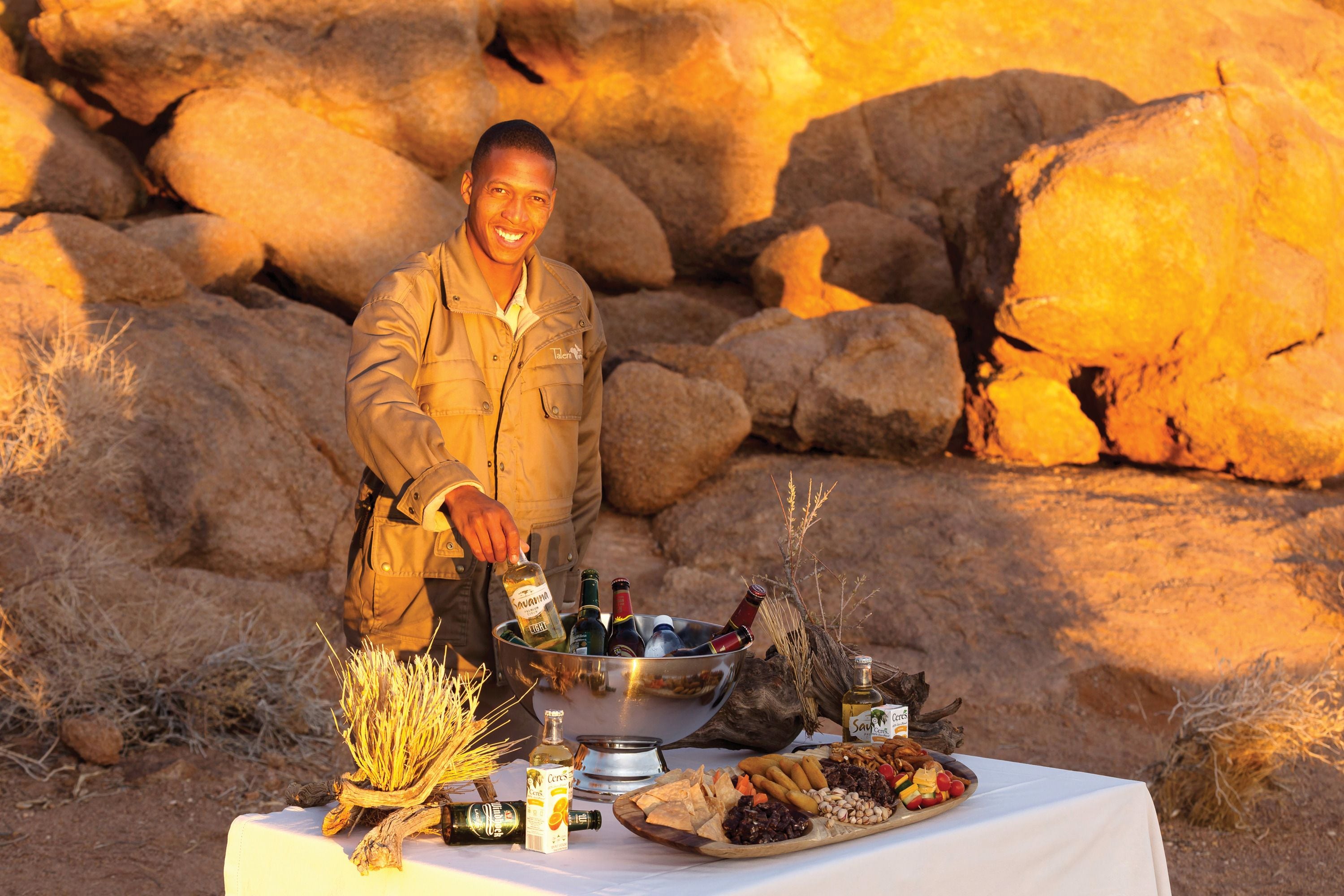 A person pouring a drink from a bottle into a metal bowl, with a table set with various food items and bottles in an outdoor setting with rocks and dry grass.