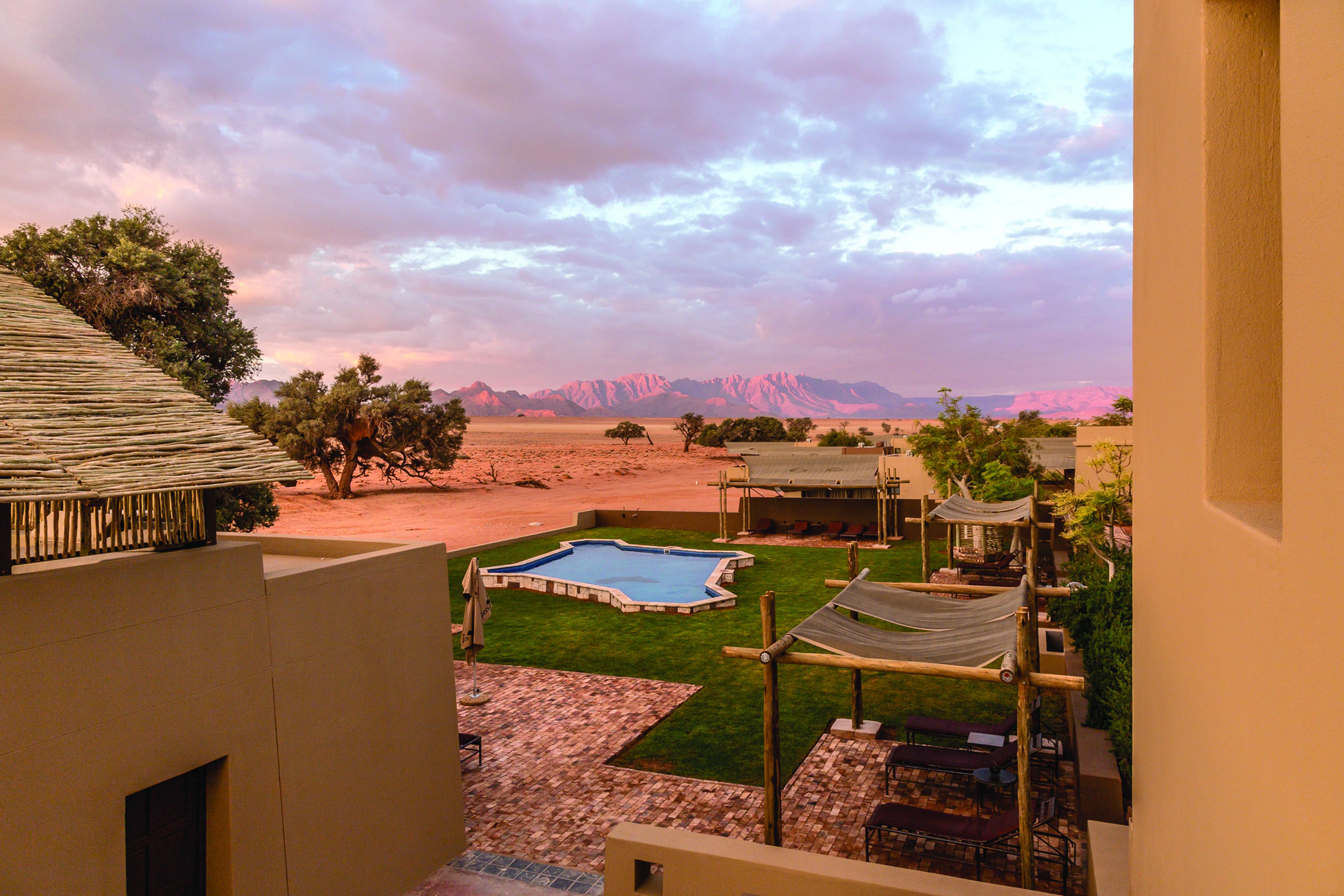 A pool area with a view of mountains, a grassy lawn, and a building with a thatched roof.