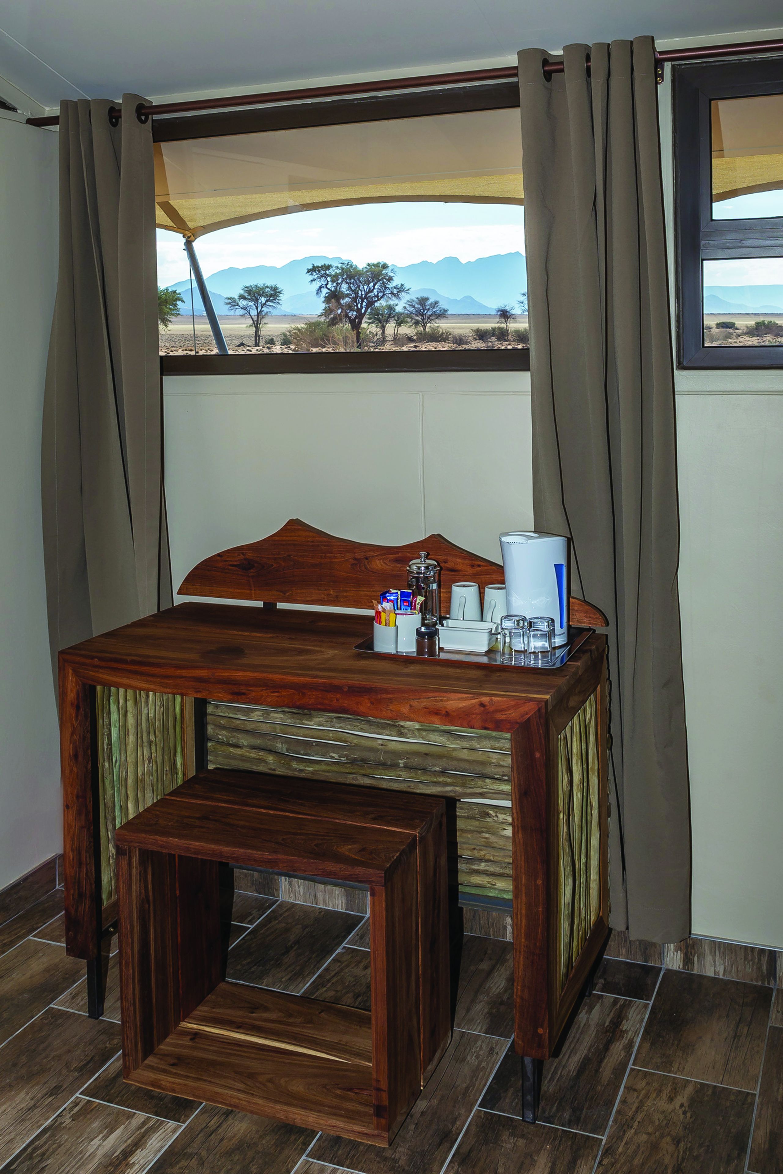 A wooden table with a coffee maker and cups, a small side table, and a view of a desert landscape through a window with curtains.
