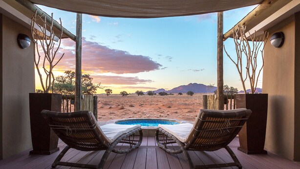 A deck with two wicker lounge chairs facing a pool and a desert landscape. - Sossusvlei Lodge (Sesriem)