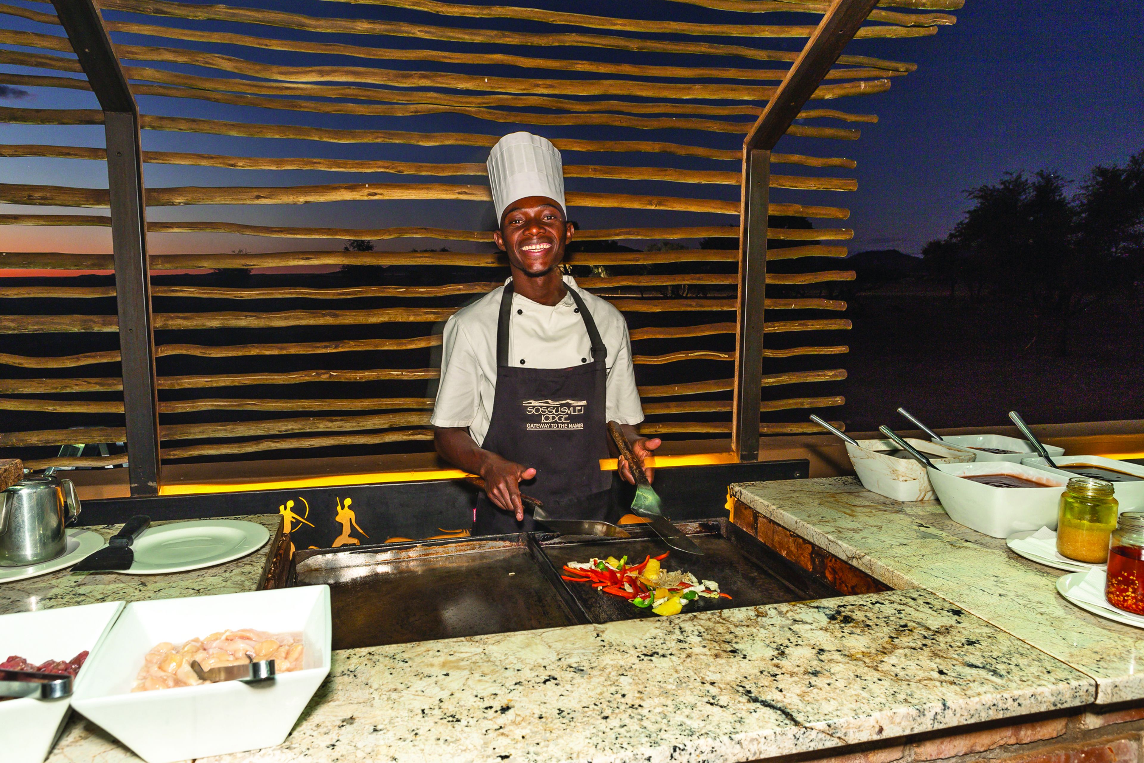 A chef at a cooking station with a grill, bowls of ingredients, and cooking utensils.