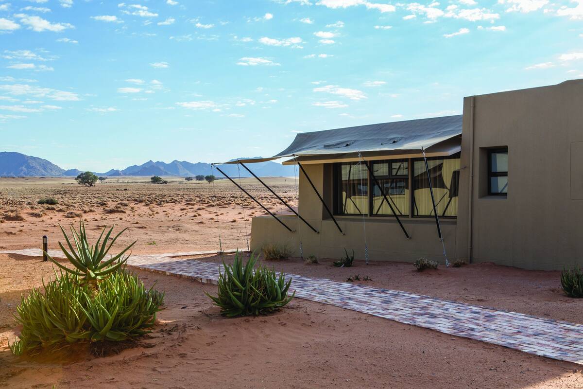 A modern building with a flat roof and large windows, surrounded by a desert landscape with sparse vegetation and distant mountains.