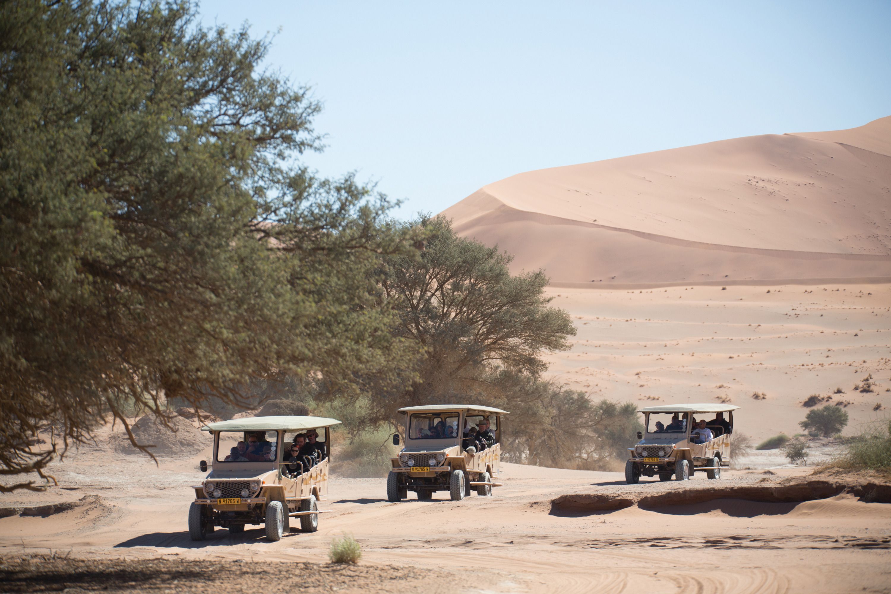 Three off-road vehicles on a sandy terrain with sparse vegetation and sand dunes in the background.