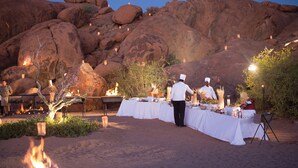 An outdoor dining setup with a chef preparing food, surrounded by large rocks and desert vegetation. - Sossusvlei Lodge (Sesriem)
