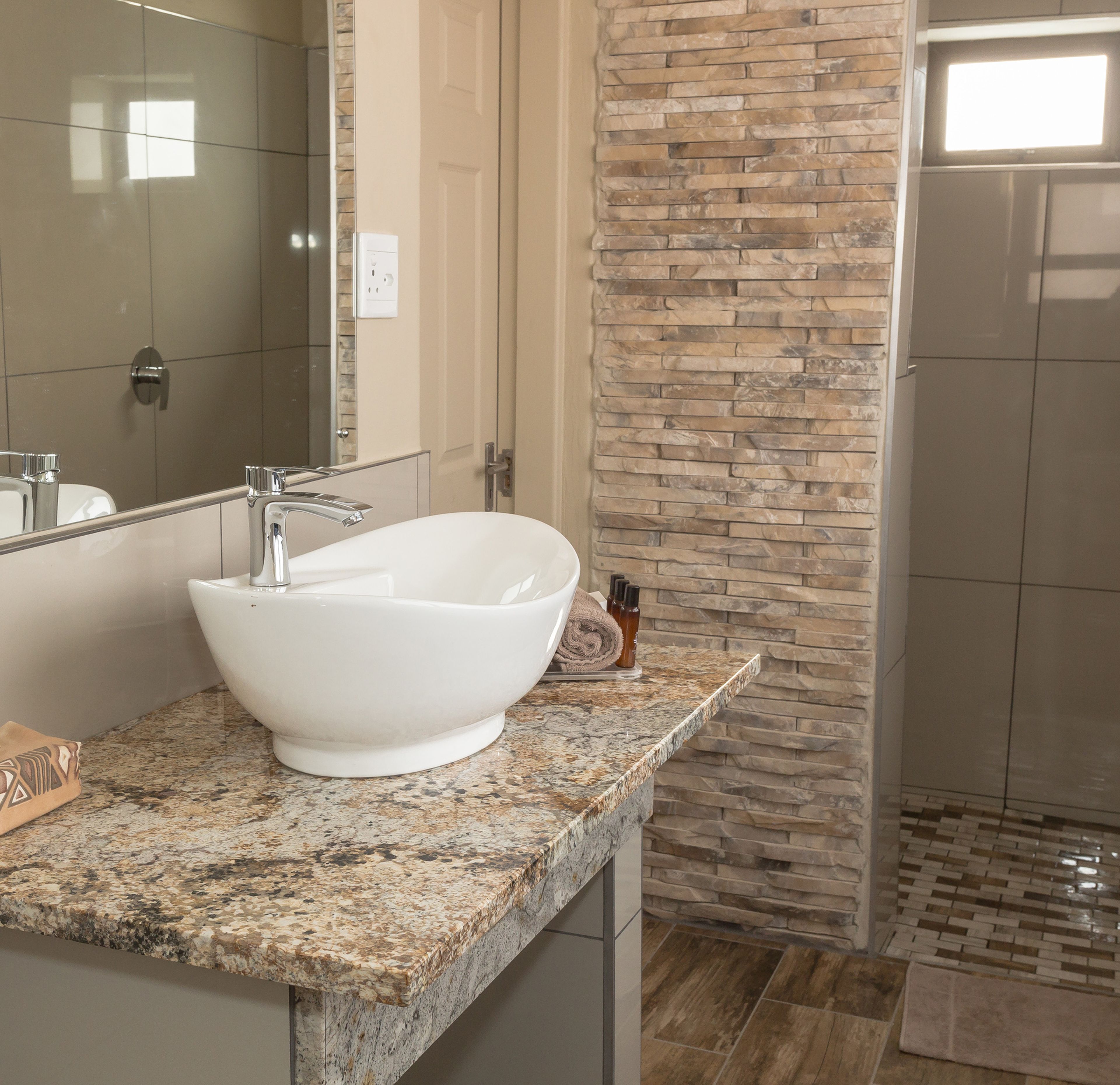 A bathroom with a white freestanding sink, a granite countertop, a stone wall, and a shower area.