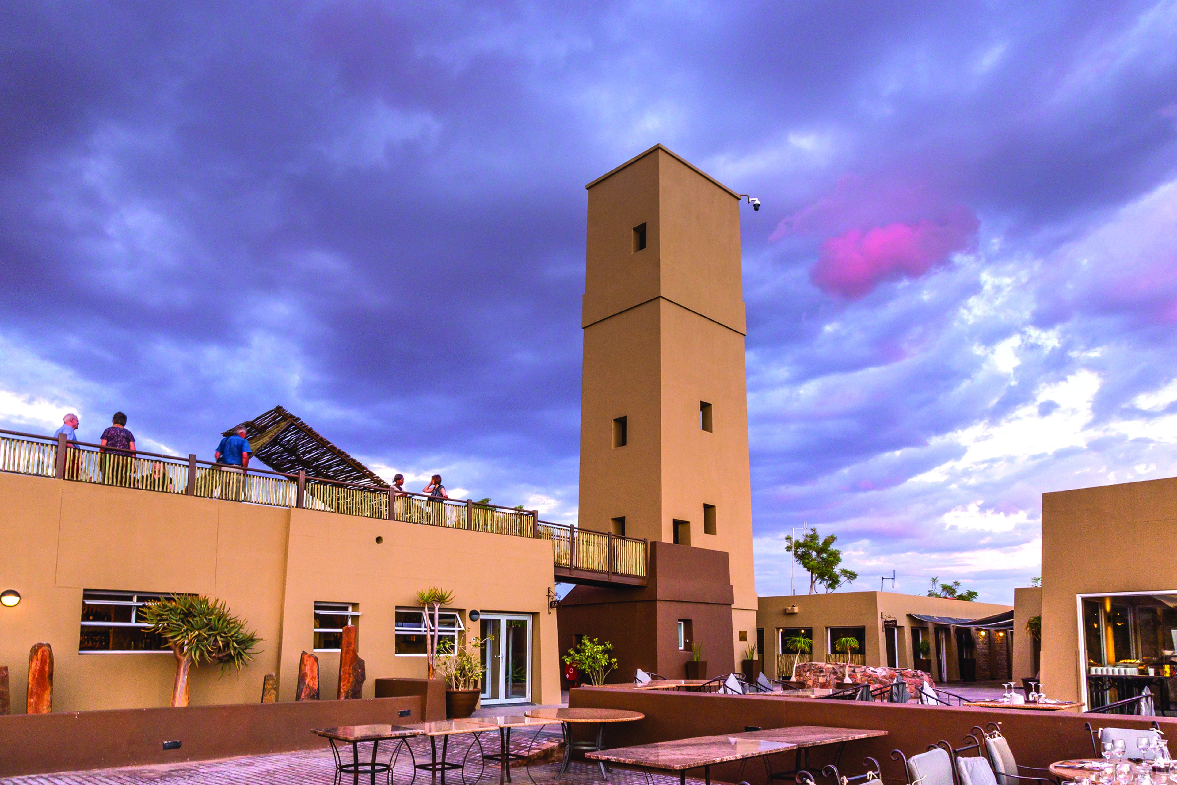 An outdoor patio area with tables and chairs, a tall tower, and a building with a balcony.