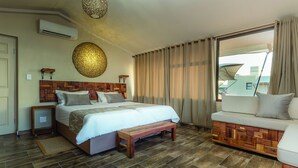 A bedroom with a wooden headboard, a large bed, a bench, a sofa, and a view through a window with curtains. - Sossusvlei Lodge (Sesriem)