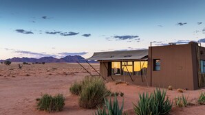 A desert landscape with a modern building featuring a large window and a shaded area. - Sossusvlei Lodge (Sesriem)