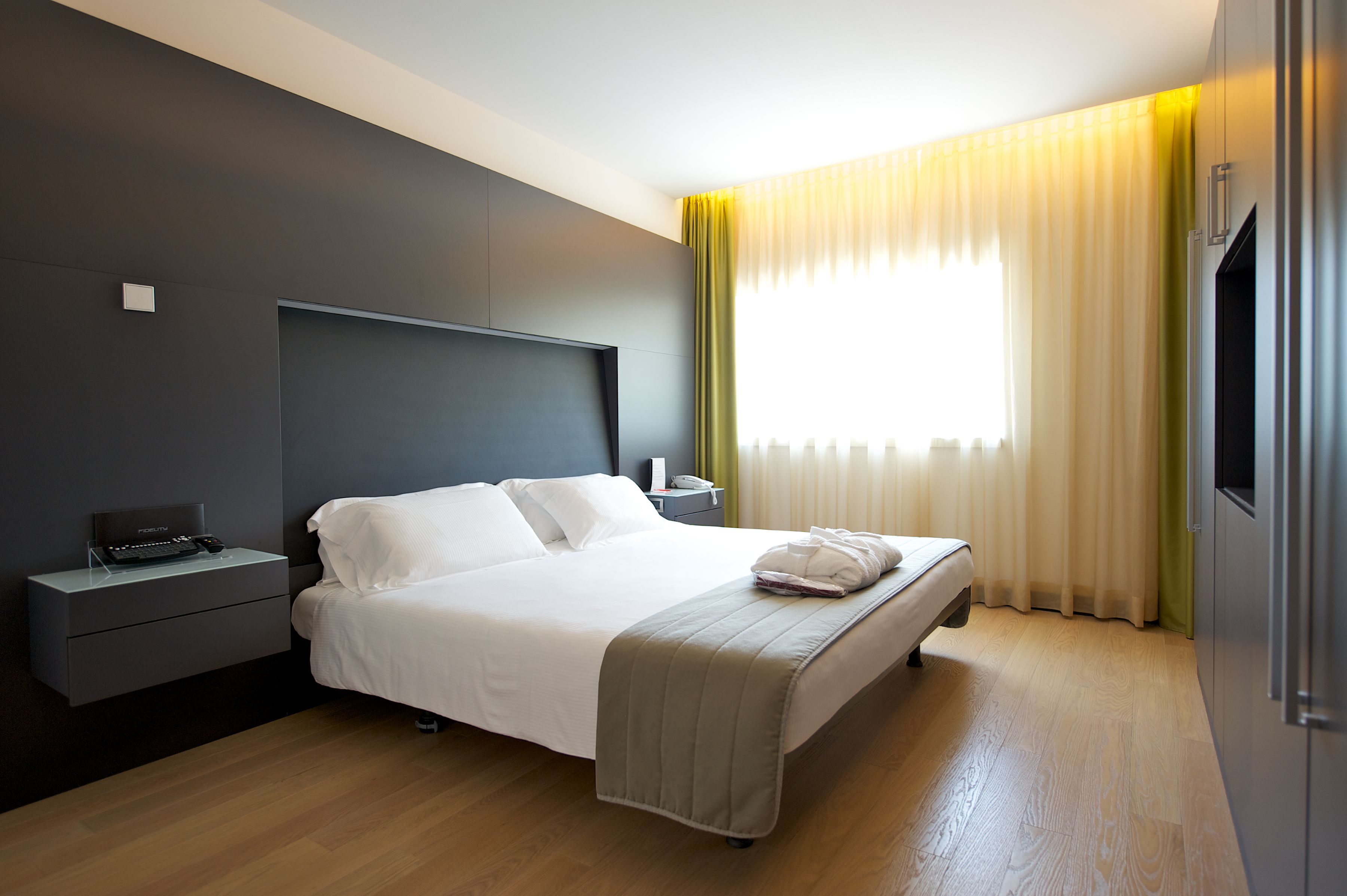 Ein modernes Hotelzimmer mit einem großen Bett, Nachttischen, einem Flachbildfernseher und einem Fenster mit Vorhängen.