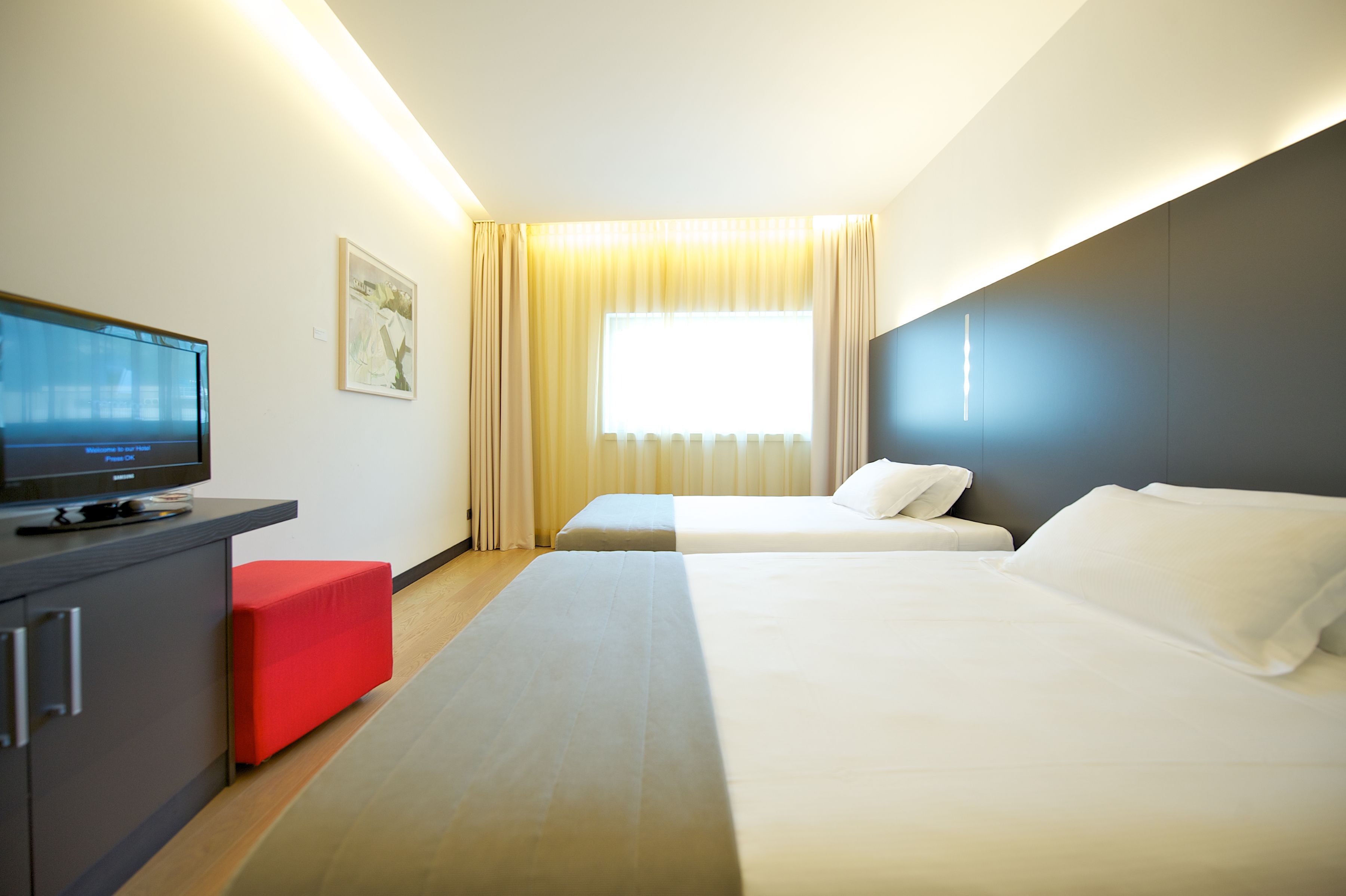 A hotel room with a bed, a TV on a stand, a red ottoman, and a window with curtains.