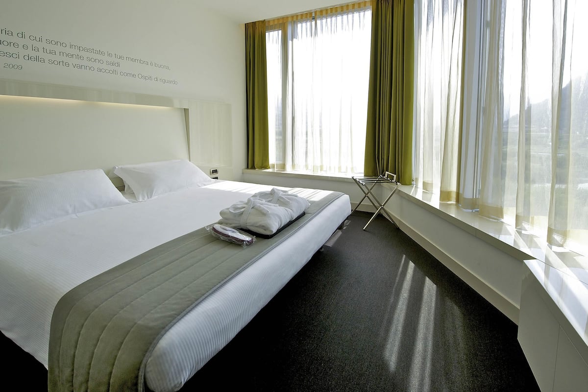A hotel room with a large bed, a window with curtains, a bedside table, and a wall-mounted light.