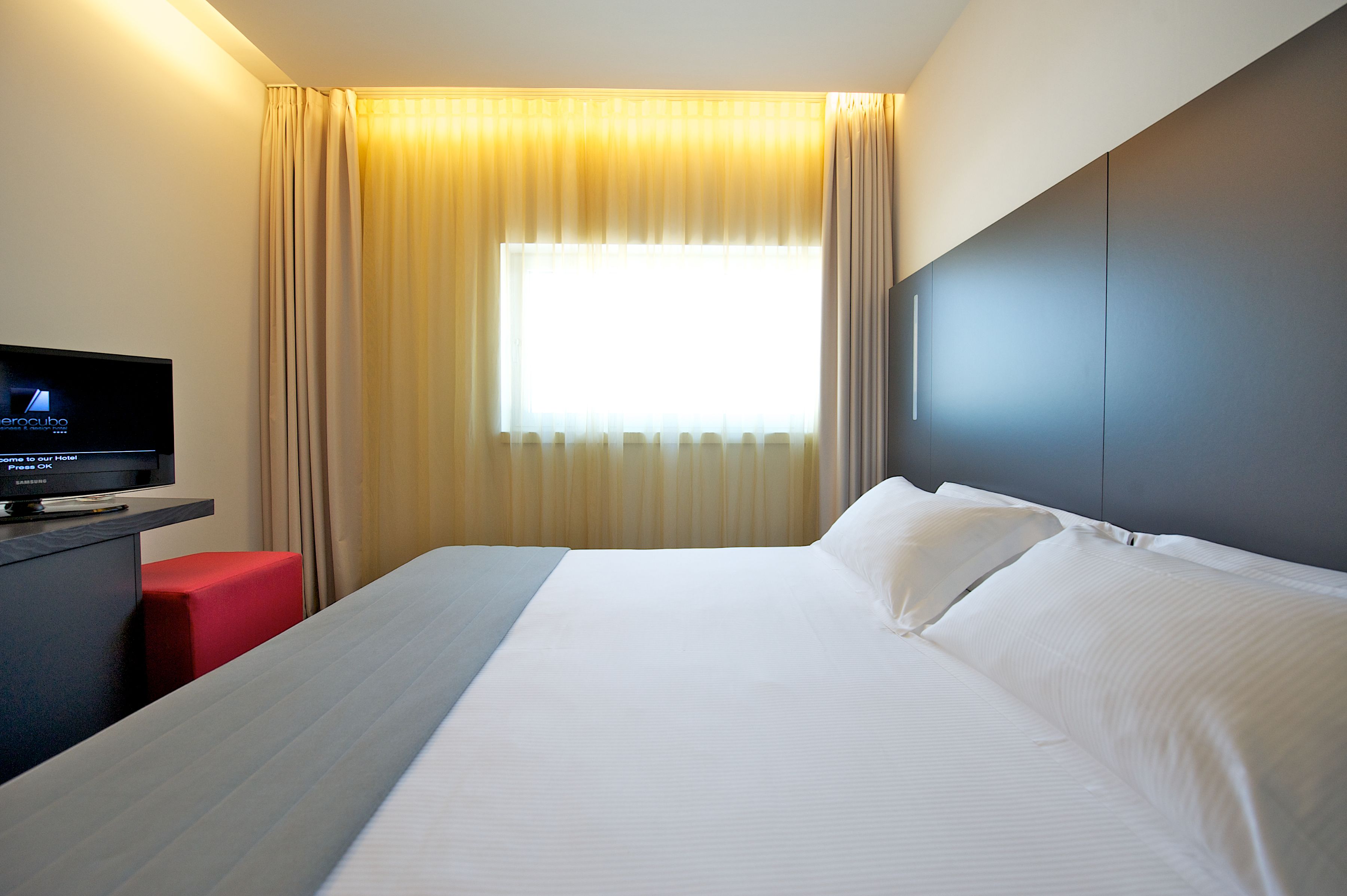 A hotel room with a large bed, a flat-screen TV, a red chair, and a window with curtains.