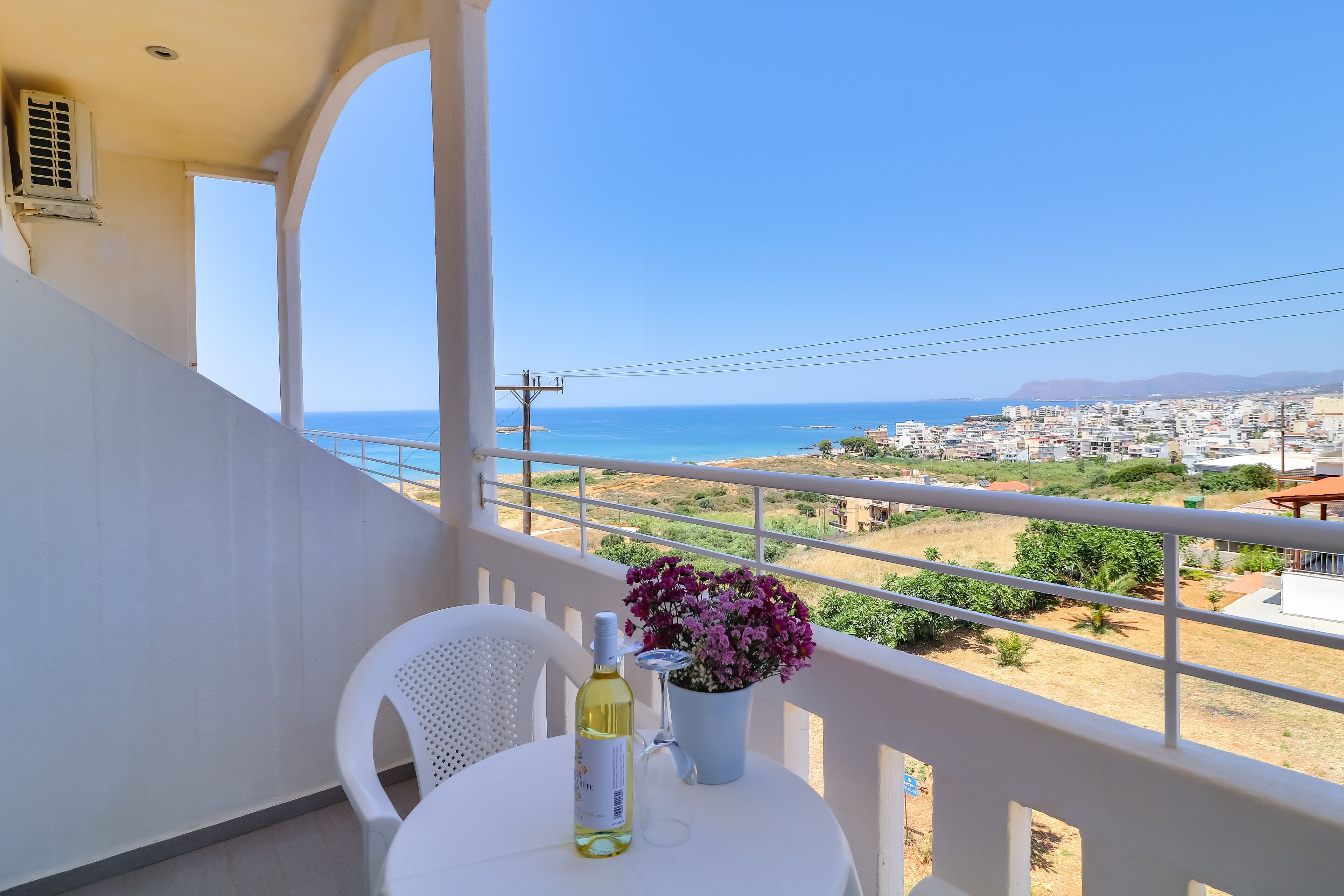 A balcony with a table, chairs, a bottle, and a flower vase overlooking a coastal town.