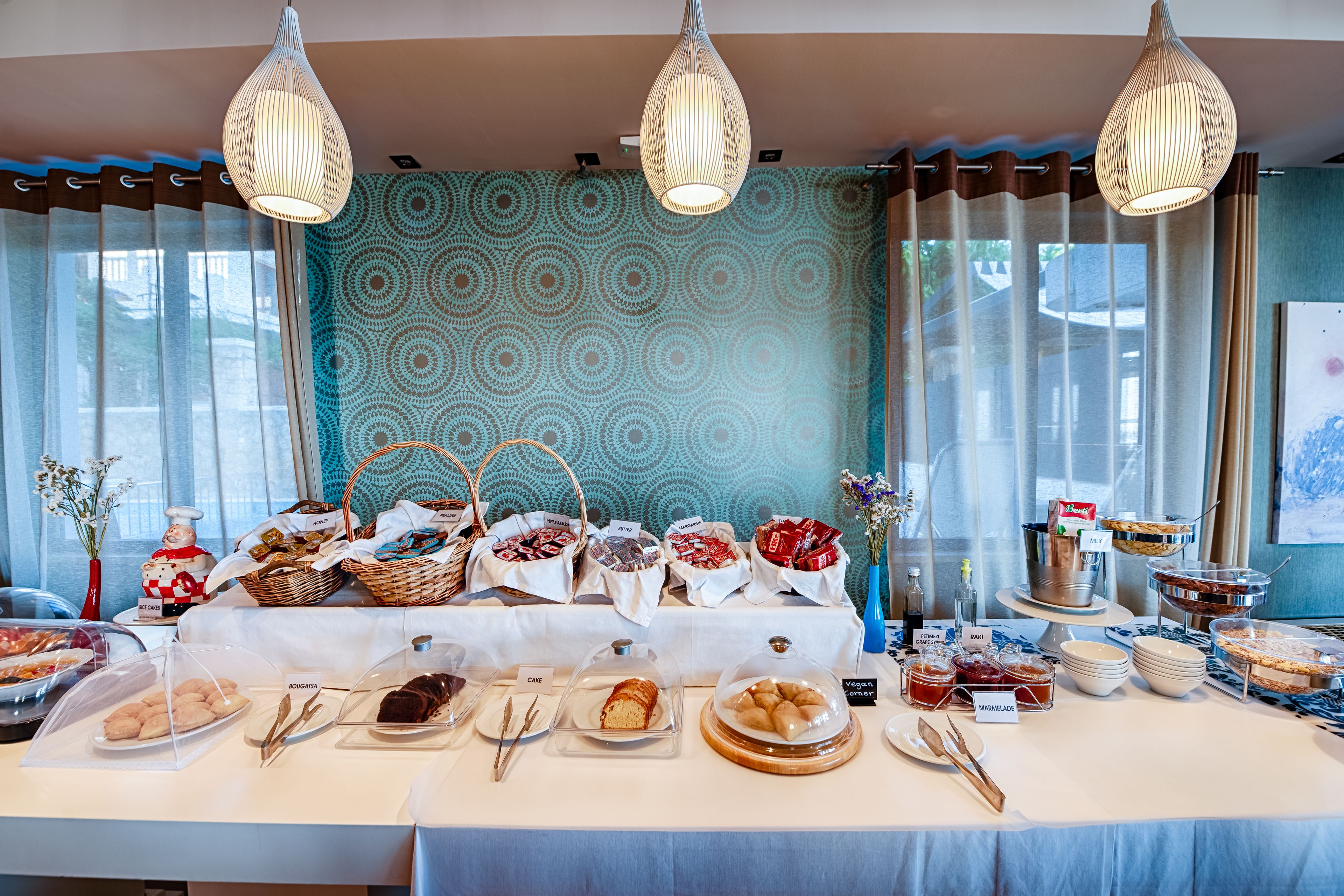 Une table de buffet garnie de divers plats et couverts, placée devant un mur à motifs et agrémentée de rideaux.