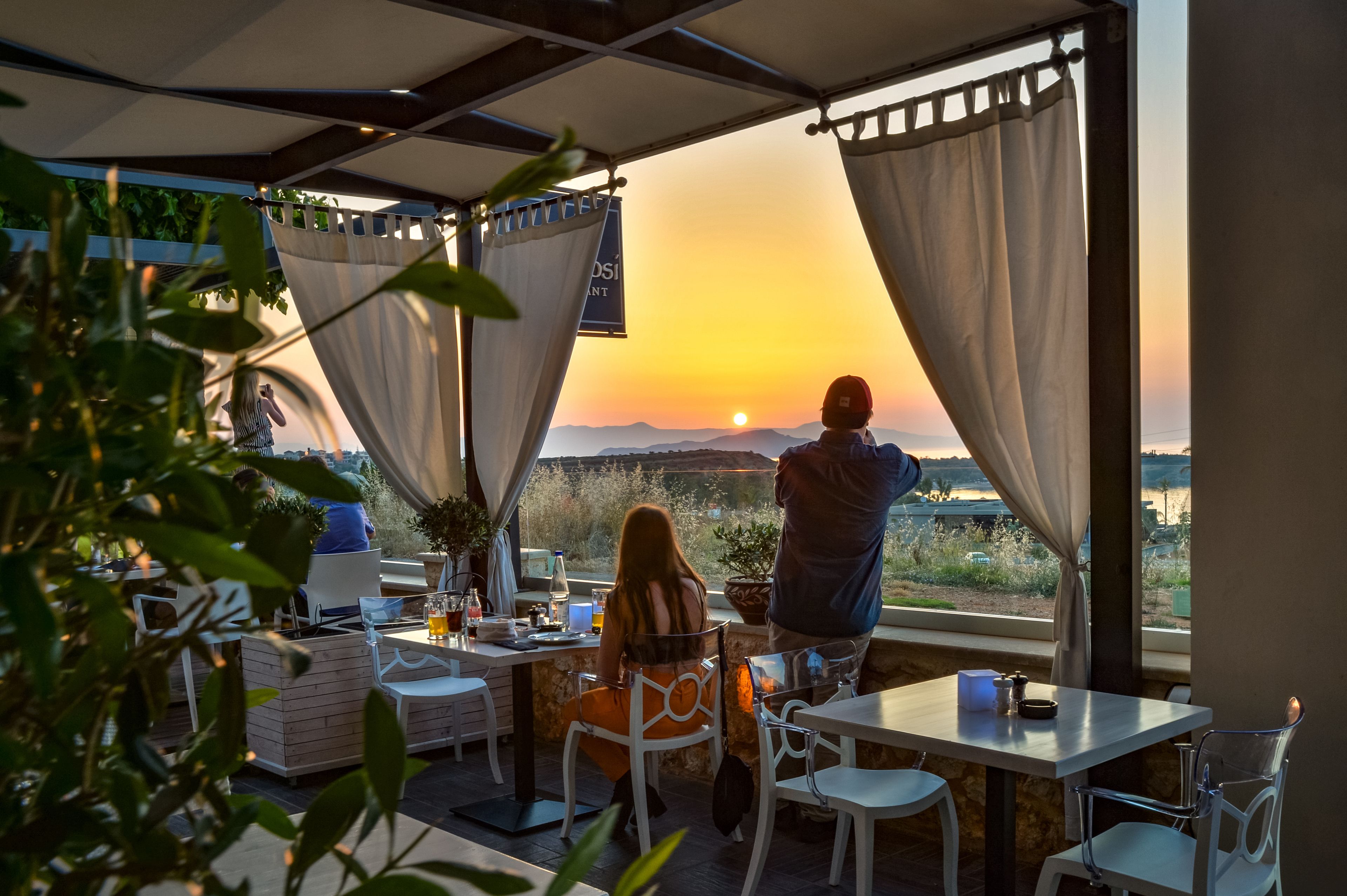 Un espacio al aire libre con mesas y sillas, una vista del atardecer y una persona de pie junto a la ventana.