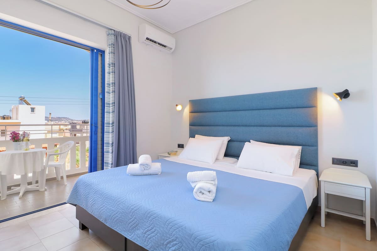 Una habitación de hotel con una cama grande, una mesita de noche, un balcón con mesa y sillas, y vistas a la ciudad.