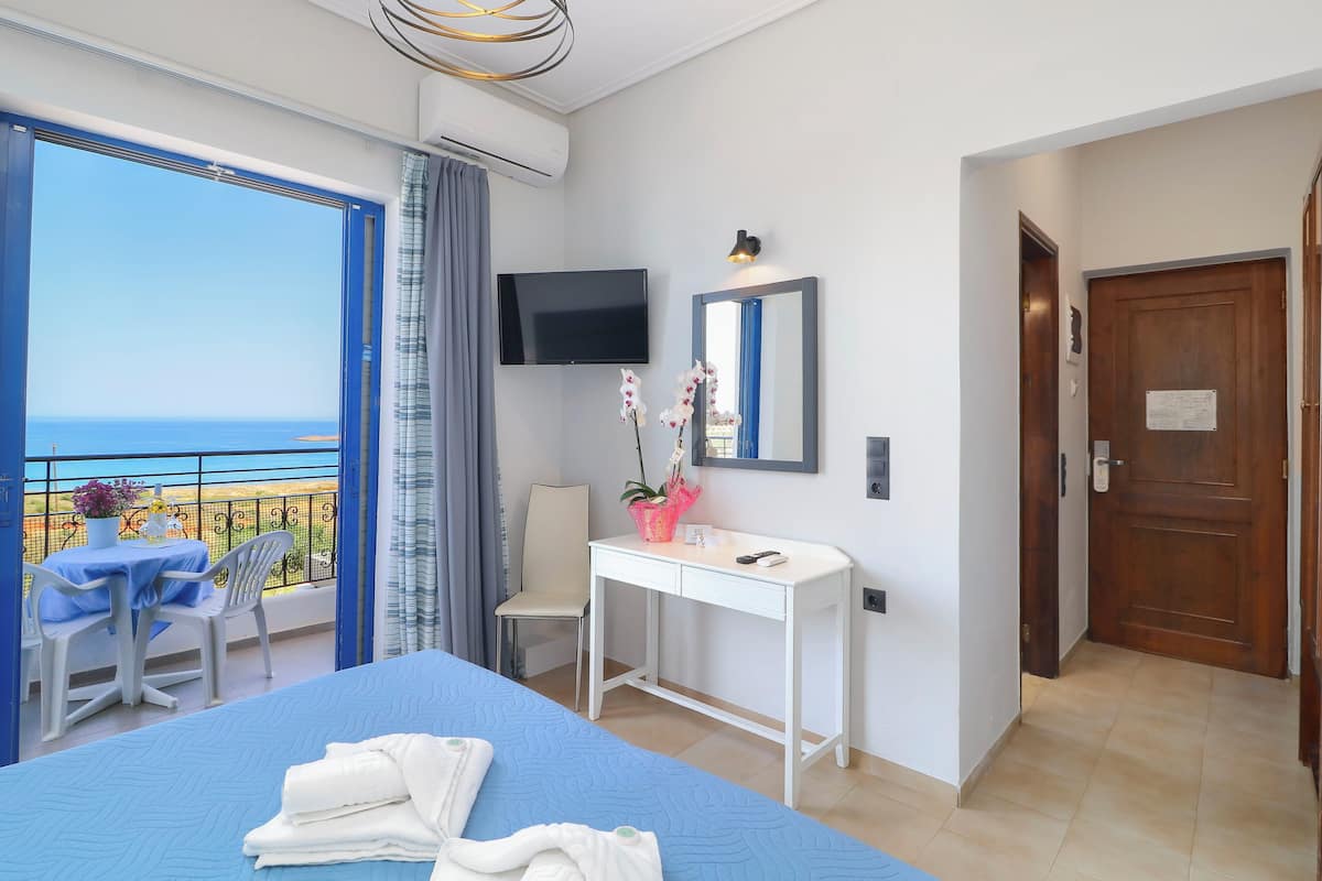 Habitación de hotel con una cama grande, un escritorio, una televisión y un balcón con vistas al mar.