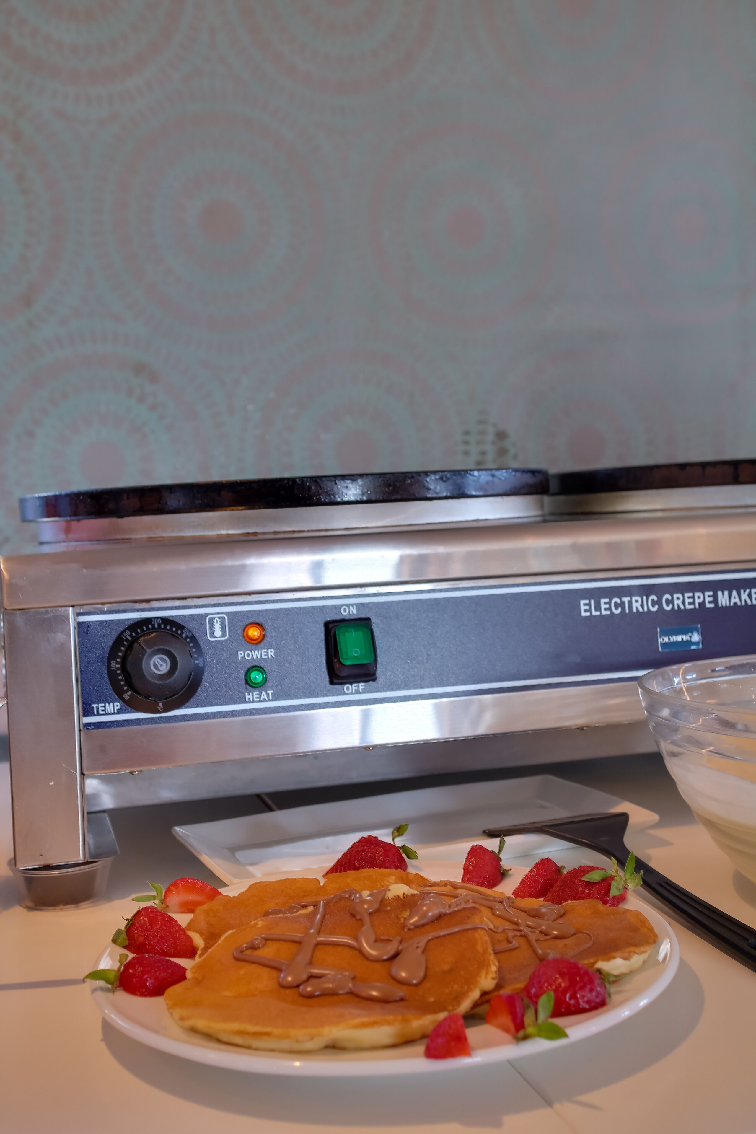 A kitchen with an electric crepe maker, a plate of crepes with strawberries and chocolate sauce, and a bowl of whipped cream.