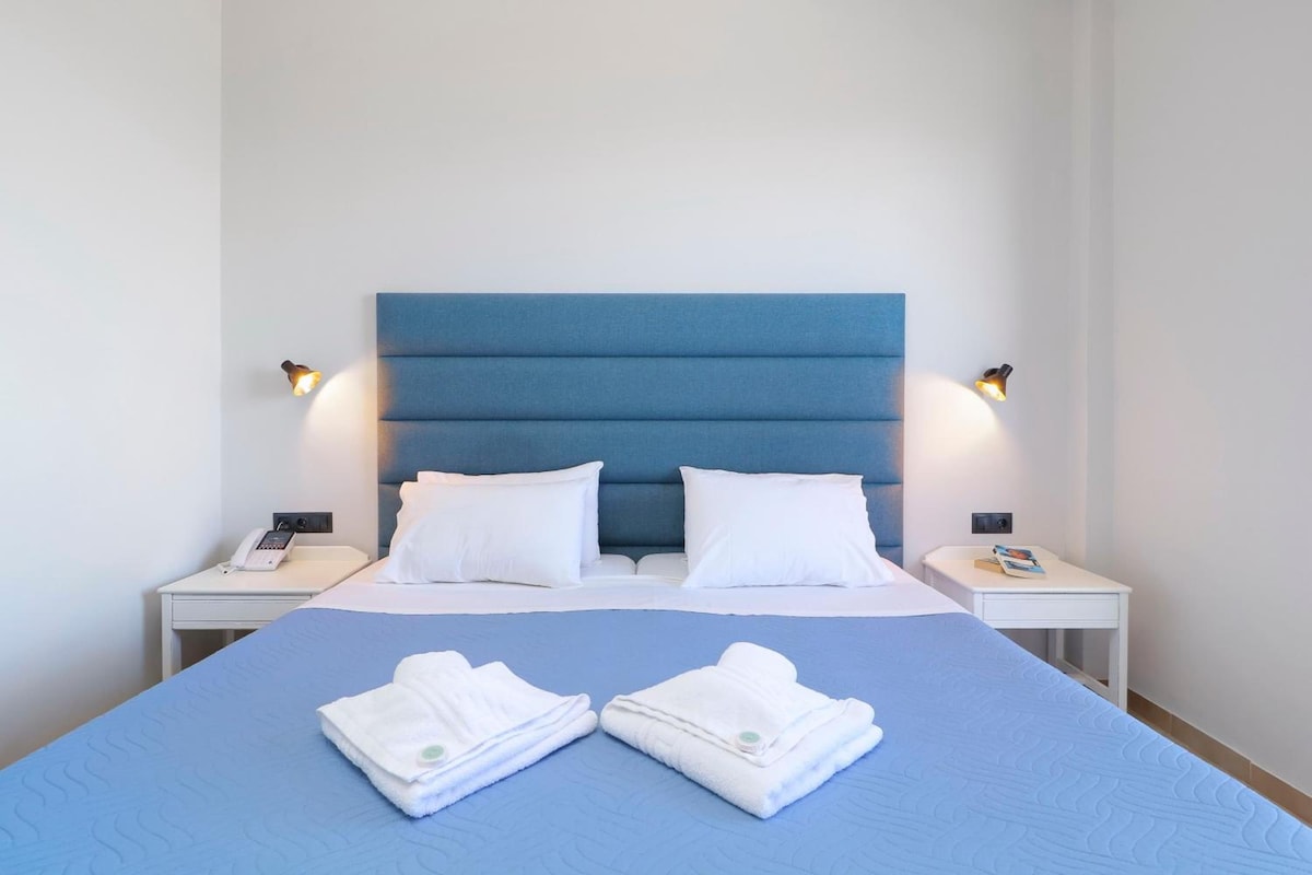 Habitación de hotel con una cama grande, dos almohadas blancas, cabecera azul, lámparas de noche y dos toallas dobladas sobre la cama.