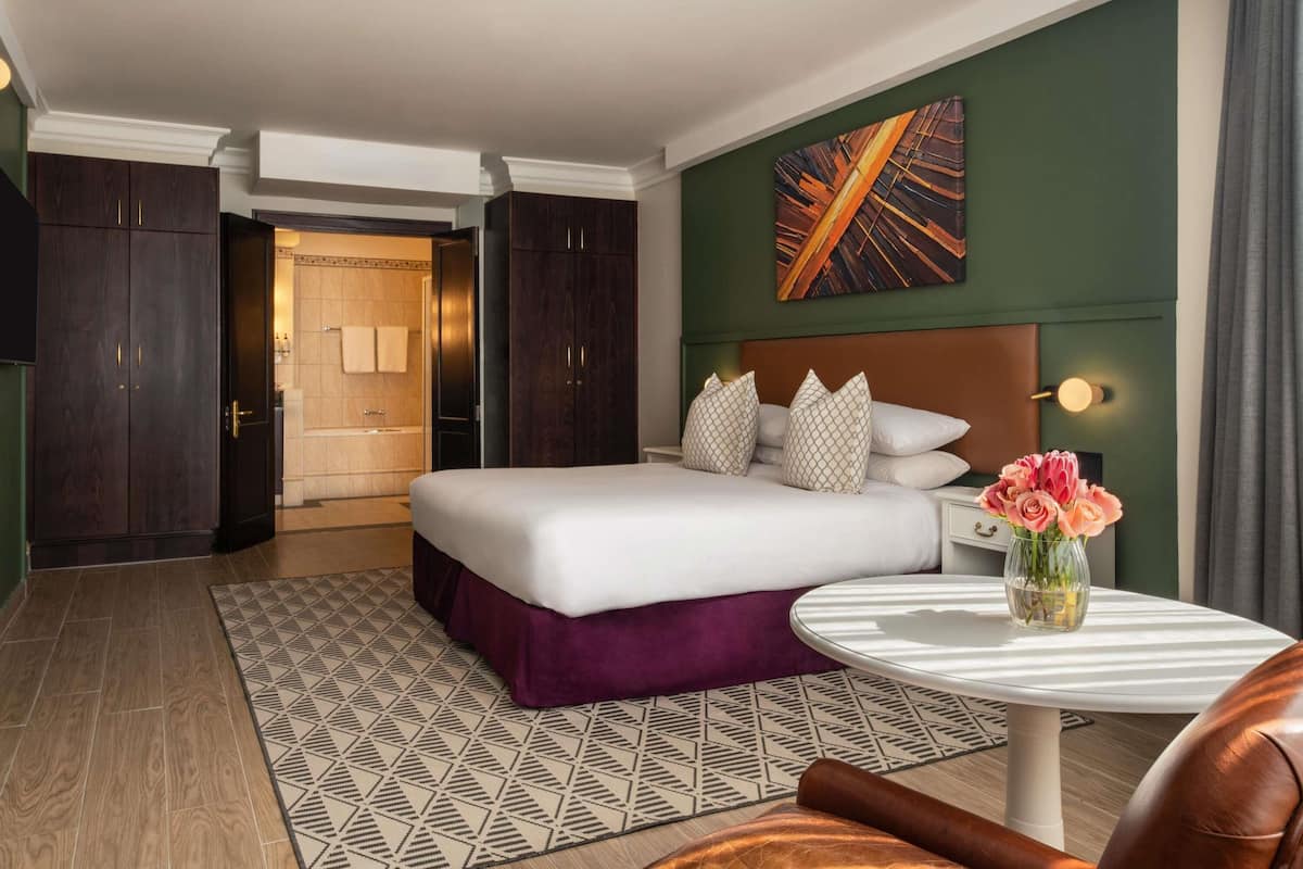 A hotel room with a bed, a round table, a vase of flowers, a painting on the wall, and a view of the bathroom.