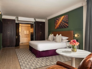 A hotel room with a bed, a round table, a vase of flowers, a painting on the wall, and a view of the bathroom.