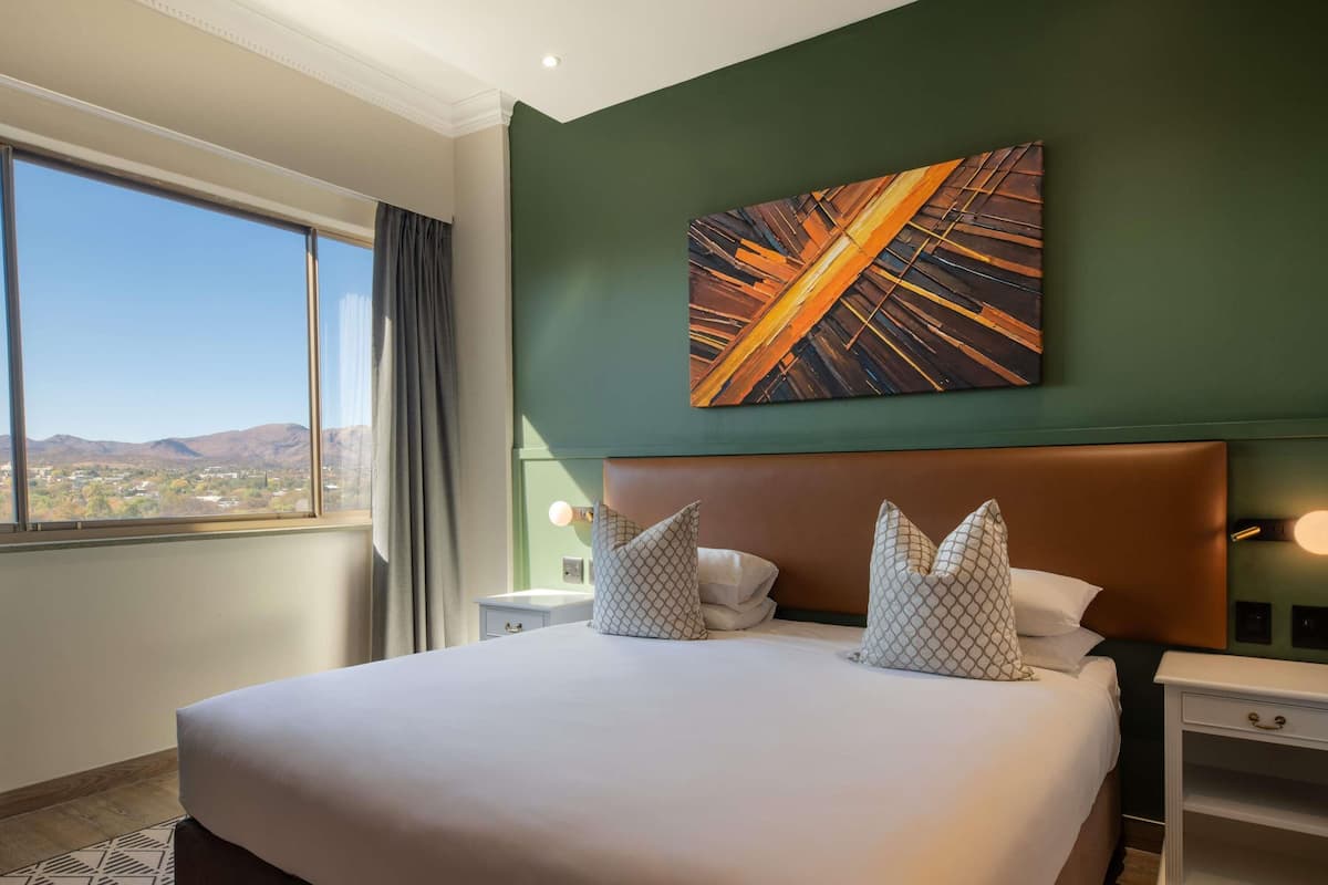 A hotel room with a large bed, a view of the city, and a painting on the wall.
