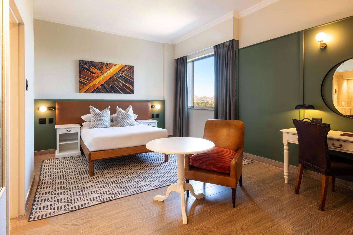 A hotel room with a bed, a desk, a chair, a small table, a rug, a painting, and a window with curtains.