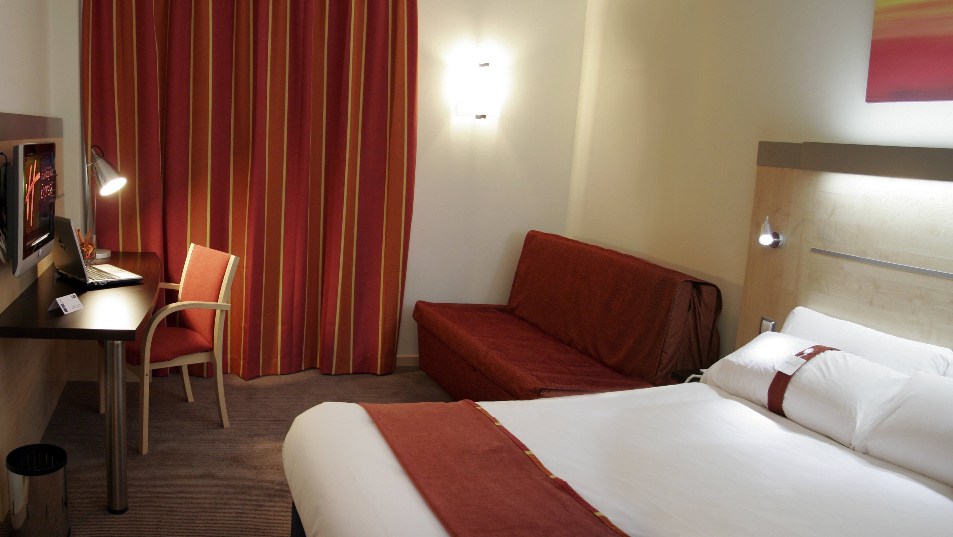 A hotel room with a bed, a desk with a laptop, a chair, and a striped curtain.